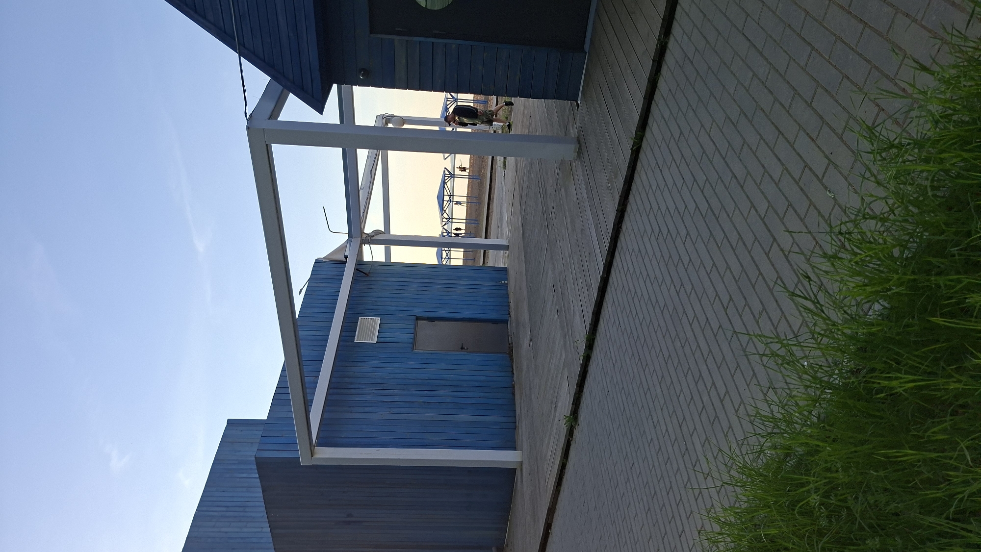 The image depicts a scene in Chaikovsky, Russia. A blue wooden structure with a door and ventilation grille is partially visible. A white metal frame surrounds part of the structure. In the background, there are beach umbrellas and a person bending over. The foreground shows a tiled walkway with grass growing along the edge. The sky is visible in the background, appearing light blue.