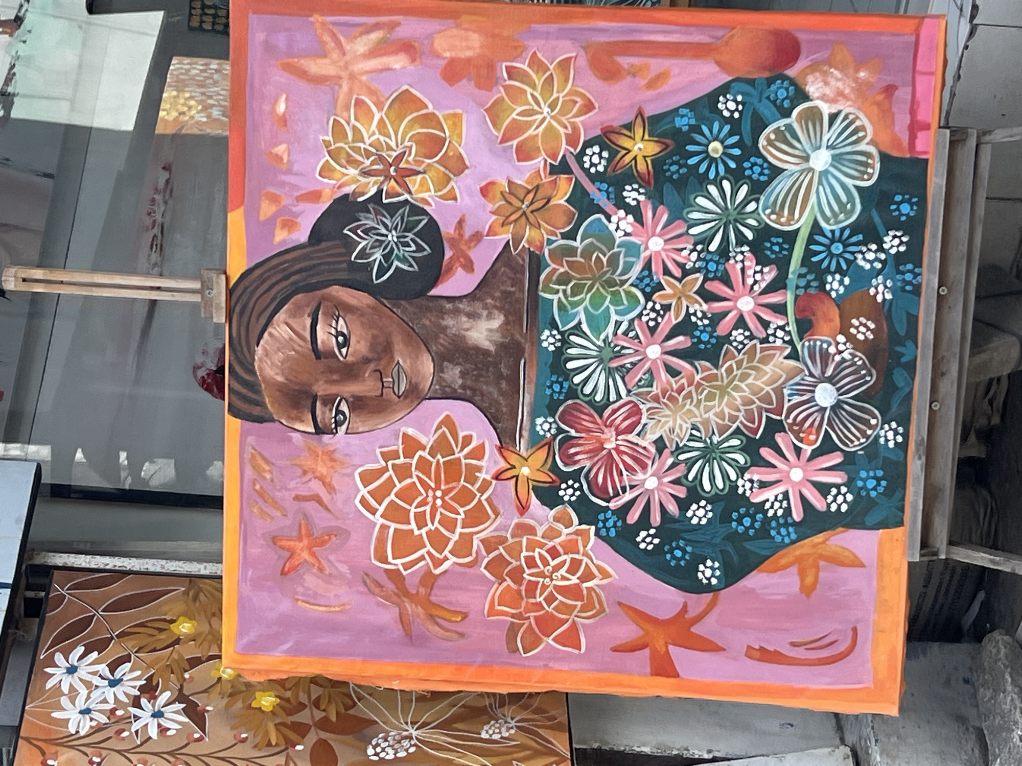 A vibrant, large-scale painting of a woman adorned with a plethora of colorful flowers is displayed on an easel outdoors, likely in a bustling urban setting like Kuta, Indonesia, indicated by the backdrop of shop fronts and displays. The painting features a dark-skinned woman with brown hair, her face rendered with striking detail, and her chest covered in an assortment of stylized floral patterns in shades of pink, red, orange, yellow, white, and blue. The background of the painting is a soft pink, with scattered orange star-like shapes and floral elements. The painting is framed by a thick, bright orange border. To the left of the main painting, another canvas is partially visible, featuring a simpler design of brown leaves and white and yellow flowers. The scene suggests an art vendor or display in a public space, with glimpses of a shop window and an outdoor environment.