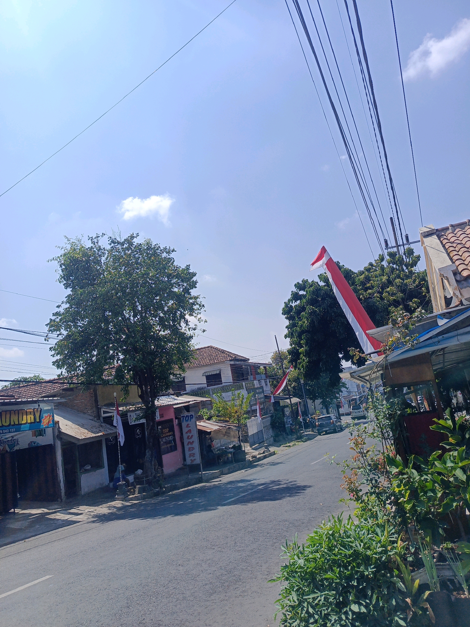 The image depicts a street scene in the city of Kepolorejo, Indonesia, under a clear blue sky with scattered clouds. On the left side of the road, several small businesses are visible, including a laundry service with a sign that reads "LAUNDRY" in blue lettering. Adjacent to it, another establishment has a pink facade and a sign indicating "TOP LAUNDRY." A tall, leafy green tree stands prominently in front of these buildings.
A red and white flag, likely the Indonesian flag, is displayed on a pole in front of one of the shops. Several overhead electrical wires crisscross the sky above the street. The road itself is paved and appears to be a two-lane street with a white line marking the center. In the distance, more buildings and trees line the street, and a few vehicles are present, including a dark-colored car. The right side of the image shows more foliage and the roof of a building with terracotta tiles. The overall impression is of a typical urban or semi-urban street on a sunny day.