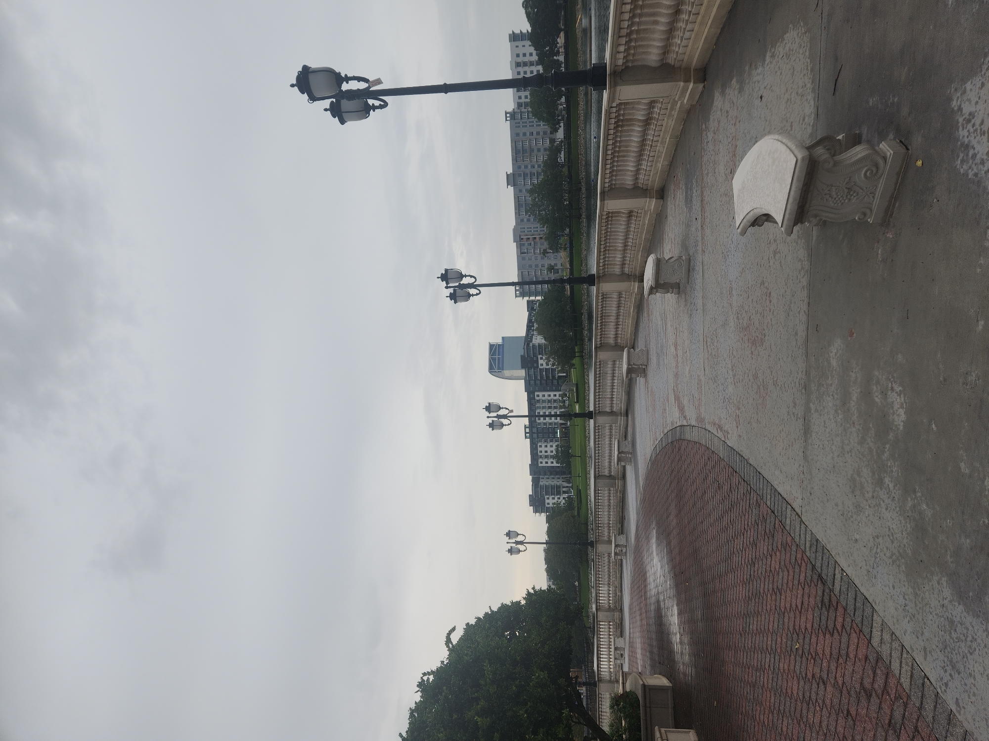 An overcast day captures a view of a waterfront promenade in Altamonte Springs, Florida. A curved pathway, paved with red bricks on the left and concrete on the right, leads the eye toward a row of ornate street lamps. Beyond the promenade, a decorative stone balustrade separates the walkway from a body of water. In the background, modern buildings with multiple stories stand against a gray sky, reflecting a cityscape. Several stone benches are strategically placed along the promenade, inviting rest. A large tree on the left adds a touch of nature to the urban landscape. The overall scene suggests a serene, yet active, public space.