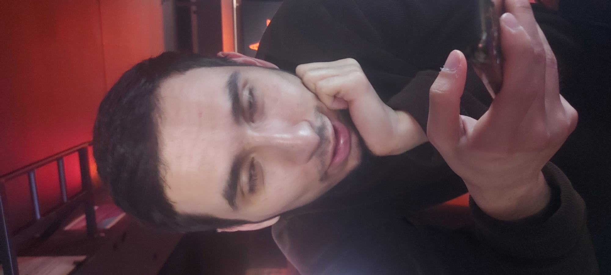 A person is looking at their phone. The individual has short dark hair, dark eyebrows, a mustache, and is wearing a dark-colored garment. Their left hand is partially visible, held up to their cheek. The individual appears to be indoors. Red lighting is visible in the background. There is a staircase behind the person. It is not possible to determine the location in Tashkent, Uzbekistan from the image provided.
