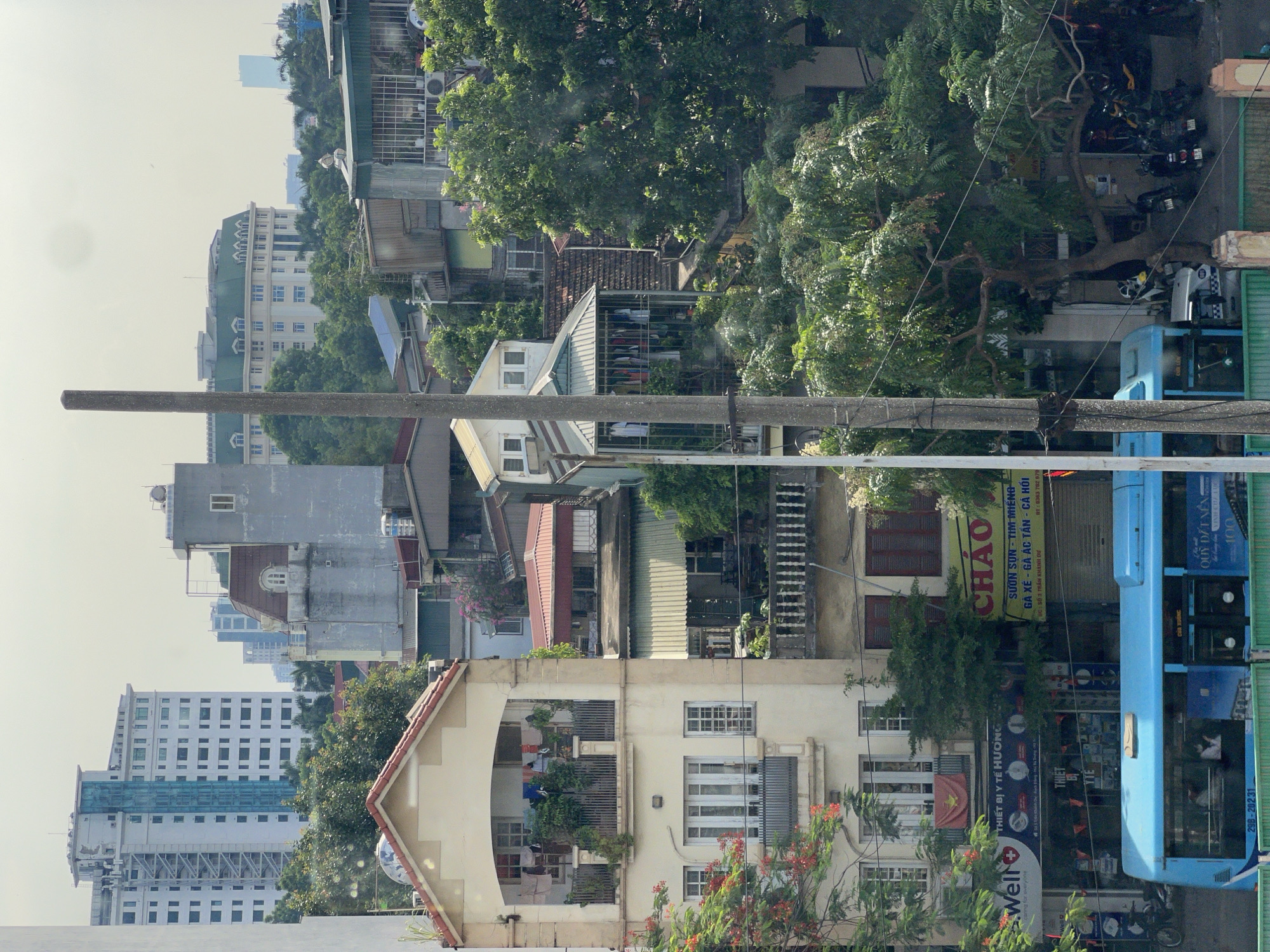 The image shows a bustling street scene in Hanoi, Vietnam, with a variety of buildings and activities. In the foreground, a bright blue bus is parked, with its doors open and passengers visible inside. The bus has the license plate "29B - 202.31". To the right of the bus, several motorcycles are parked, suggesting a common mode of transportation in the city.
The buildings lining the street display a mix of architectural styles, indicative of urban development and historical layers. A tall, cream-colored building with multiple balconies filled with plants and laundry stands prominently on the left. Above its balconies, there are arched openings with railings. Further up, the building has a red-tiled roof. To its right, another building with a more modern facade and a green roof is visible, featuring a series of windows.
In the background, a series of taller buildings, including a white high-rise with a blue glass section, dominate the skyline. The overall impression is one of a dense urban environment characterized by a lively street life, a blend of residential and commercial structures, and a common presence of greenery and trees interspersed within the city. The sky is overcast, suggesting a typical weather condition for the region.