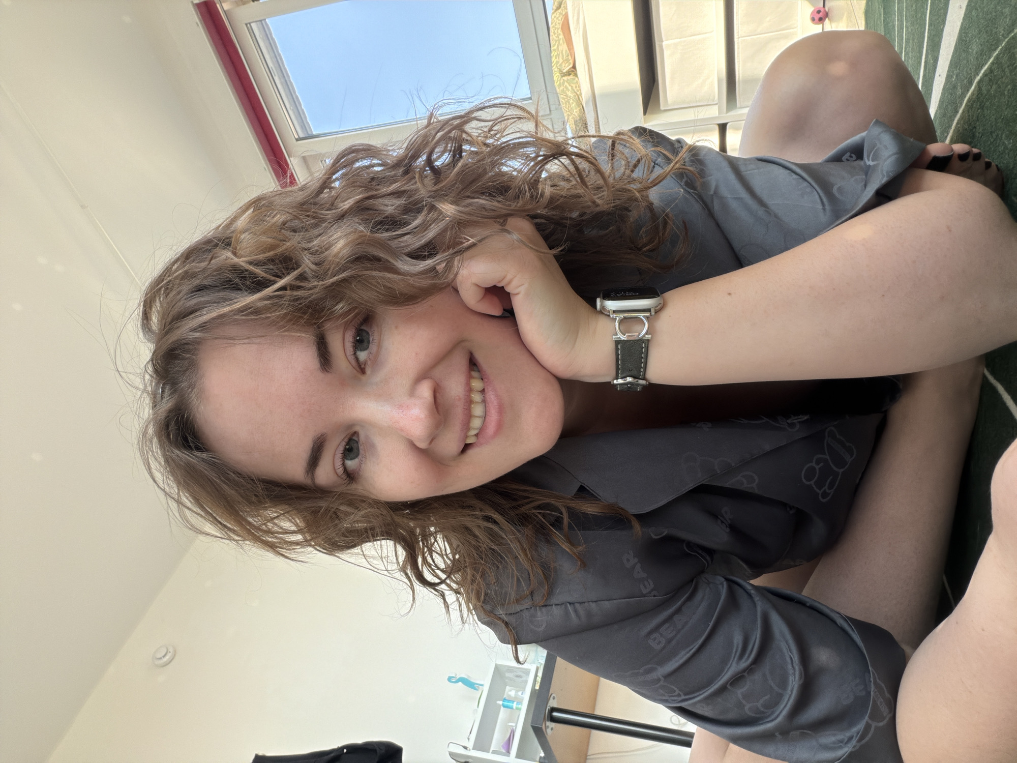 A woman with curly brown hair and fair skin is lying on her side, smiling at the camera. She is wearing a gray silky pajama top with a teddy bear pattern and a wristwatch with a black leather strap. She is resting her head on her hand. In the background, there is a window with blue sky visible, a white wall, a small shelf, a green and white striped surface, and a glimpse of bare legs. The image appears to be taken indoors. The location is Unknown, Unknown.