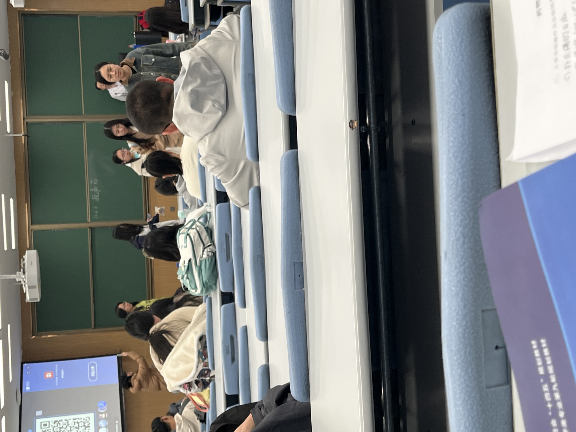 A medium, eye-level shot, taken indoors, depicts a classroom in Changchun City, China, filled with students seated at rows of desks with blue padded chairs. The classroom has a green chalkboard and an interactive display screen showing a QR code, suggesting it might be a lecture hall at a university. The students are of varying heights and wear casual clothing. In the foreground, one student is wearing a white hoodie. Papers are on the desk. The overall atmosphere suggests a learning environment.