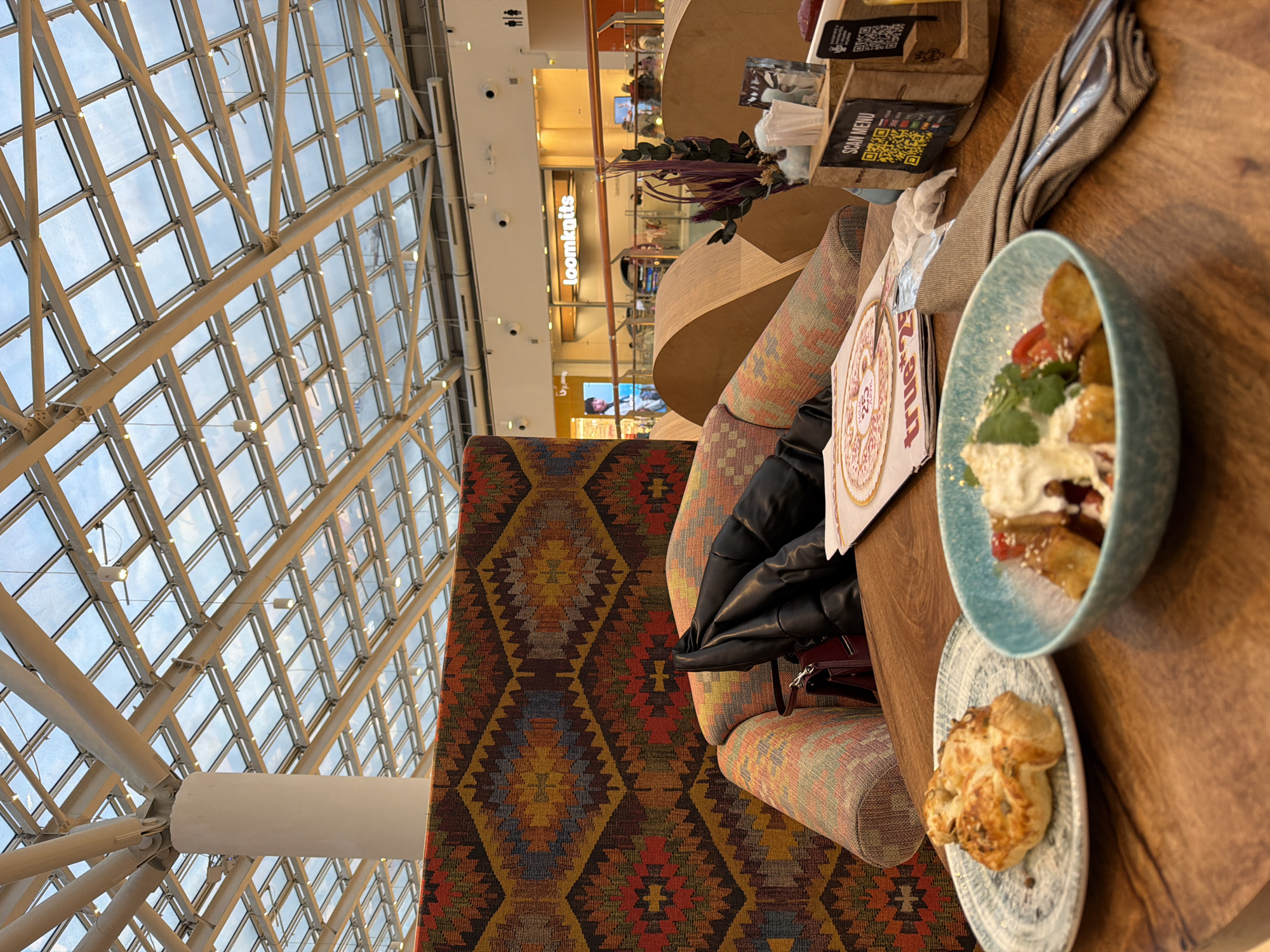 The image presents a vibrant and modern indoor scene, likely a cafe or restaurant within a large shopping mall in Khimki, Russia. The perspective is from a seated position, looking across a wooden table towards a distinctive seating area and retail shops beyond.
In the immediate foreground, a wooden table holds a light blue bowl containing a dessert or fruit salad, garnished with white cream, red berries, and what appear to be golden-brown fried or baked pieces. A smaller, patterned plate next to it features a golden pastry, possibly a scone. A black puffer jacket is draped over a chair, with a small dark red bag visible below it. A folded light brown napkin, a menu stand displaying "SCAN MENU" and QR codes, and a partially obscured flyer for "true 23" or "true 2.0" with a "25" logo are also on the table, alongside a decorative plant with purple leaves.
The seating area features a long, low sofa or bench upholstered in a striking, colorful fabric with a geometric pattern, reminiscent of an ethnic rug in shades of red, orange, yellow, and blue. An adjacent chair has a lighter, striped upholstery.
The background is dominated by a vast glass ceiling structure supported by white metal beams, letting in abundant natural light and revealing the sky, indicating daytime. Beyond the seating, multiple shops line the upper level of the mall. A prominent orange sign for "loomknits" is clearly visible. Other shops feature large display screens, and faint figures of people can be seen moving in the distance. Recessed lighting fixtures and security cameras are integrated into the modern architecture. The overall scene suggests a relaxed and casual dining experience within a bustling commercial complex.