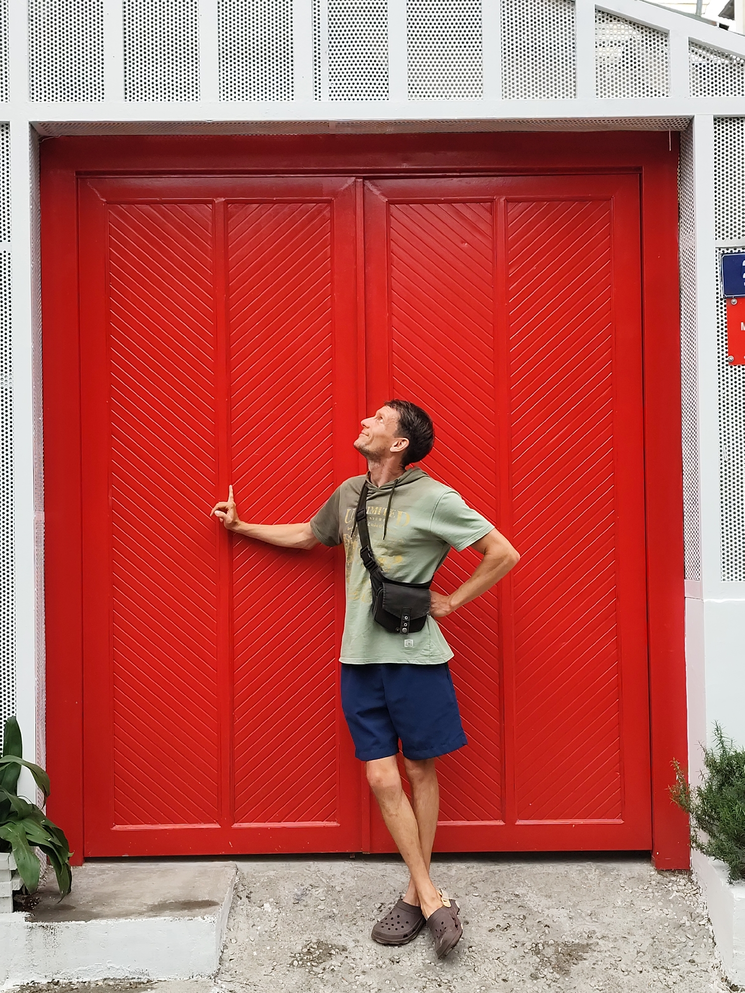 A man stands in front of a large, bright red double door, which is flanked by sections of white, perforated metal. The man is light-skinned with short dark hair. He wears a light green t-shirt, blue shorts, and brown Crocs. A black shoulder bag is slung across his chest. He is standing on a concrete surface and has his left hand resting on the left door and his right hand on his hip. He is looking upwards. The scene includes small plants to the left and right of the door. The location is Da Lat, Vietnam.