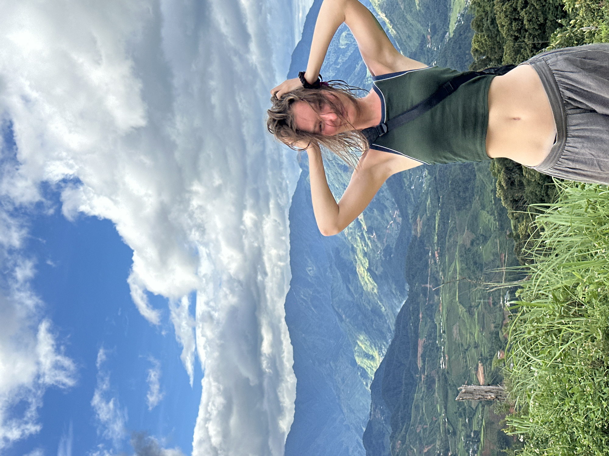 A young, light-skinned woman with brown, wind-blown hair is positioned in the foreground, smiling or laughing with her eyes squinted, her hands running through her hair. She wears a dark green sleeveless crop top with white trim and loose-fitting grey pants, exposing her midriff. A black strap, likely from a bag, crosses her chest, and a dark hair tie is visible on her left wrist. The scene is set on a high vantage point overlooking a vast mountainous landscape in the city of Hanh Phuc, Vietnam. The background features multiple layers of green and blue mountains, some appearing terraced with patches of lighter green, and scattered small buildings are visible. The sky above is partly cloudy, with both bright blue patches and large, fluffy white and grey clouds. In the immediate foreground, lush green grass and small white flowers are visible, along with a prominent weathered tree stump or trunk on the left.