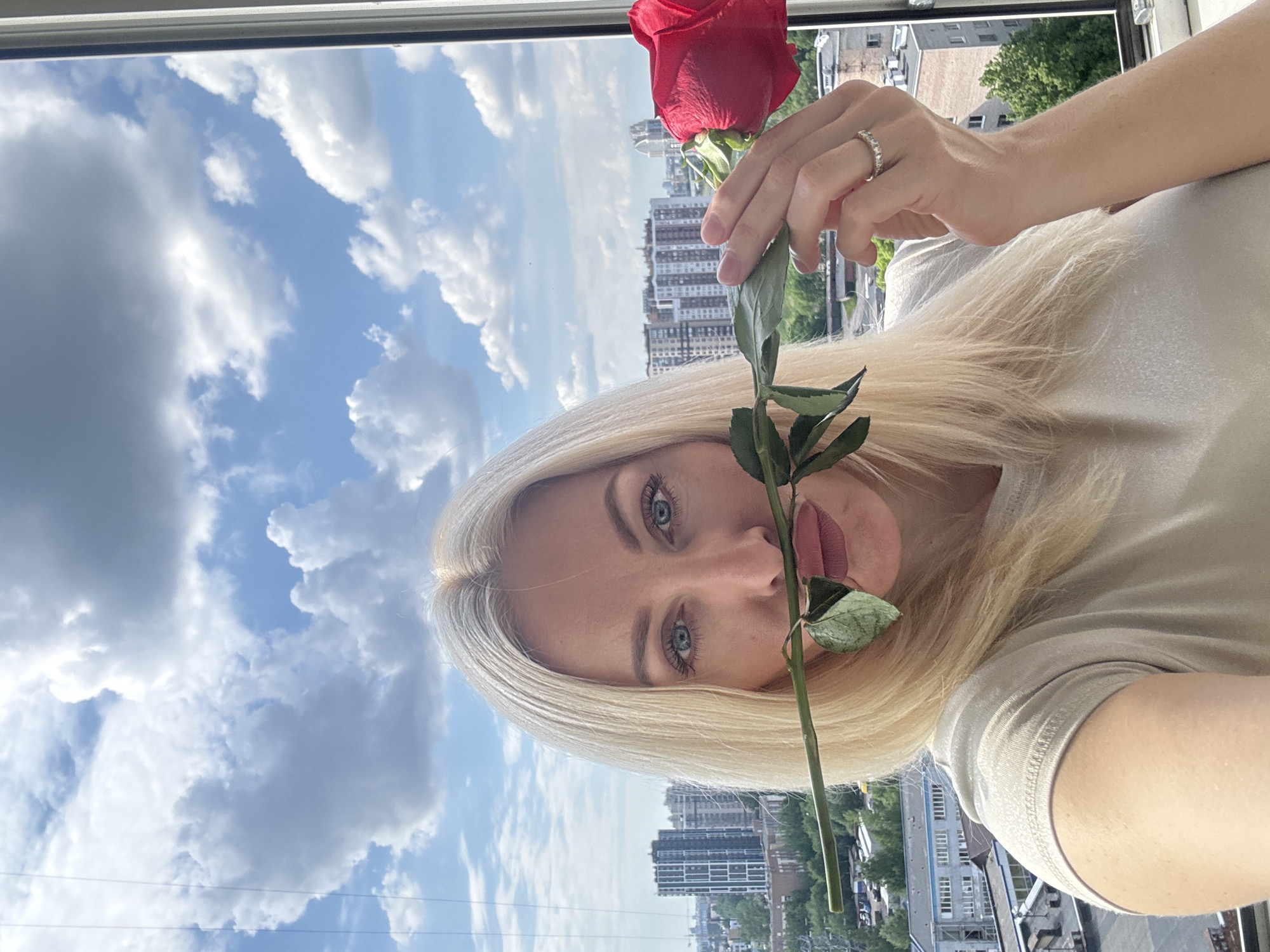 This media file is an upright portrait-style image, rotated 90 degrees counter-clockwise, featuring a person holding a red rose. The subject is a person with long blonde hair and striking blue eyes, looking directly at the viewer. A single red rose is held in their right hand, with its stem and leaves strategically placed to cover their mouth and nose. The person is wearing a light, neutral-colored top, possibly a t-shirt, and a ring with several small, clear stones is visible on their right ring finger.
The background shows a bright, daytime outdoor scene. Above the person's head (to the left in the rotated image) is a clear blue sky filled with numerous white and some darker grey cumulus clouds. Below the horizon line, a sprawling city landscape is visible, characterized by multiple multi-story residential or commercial buildings. Some green trees and foliage are also interspersed within the urban environment. The image appears to be taken from a window or balcony, with a white frame or railing visible at the top edge of the original, unrotated image. The lighting is natural and bright, suggesting a sunny day with some cloud cover. There is no visible text in the image.
