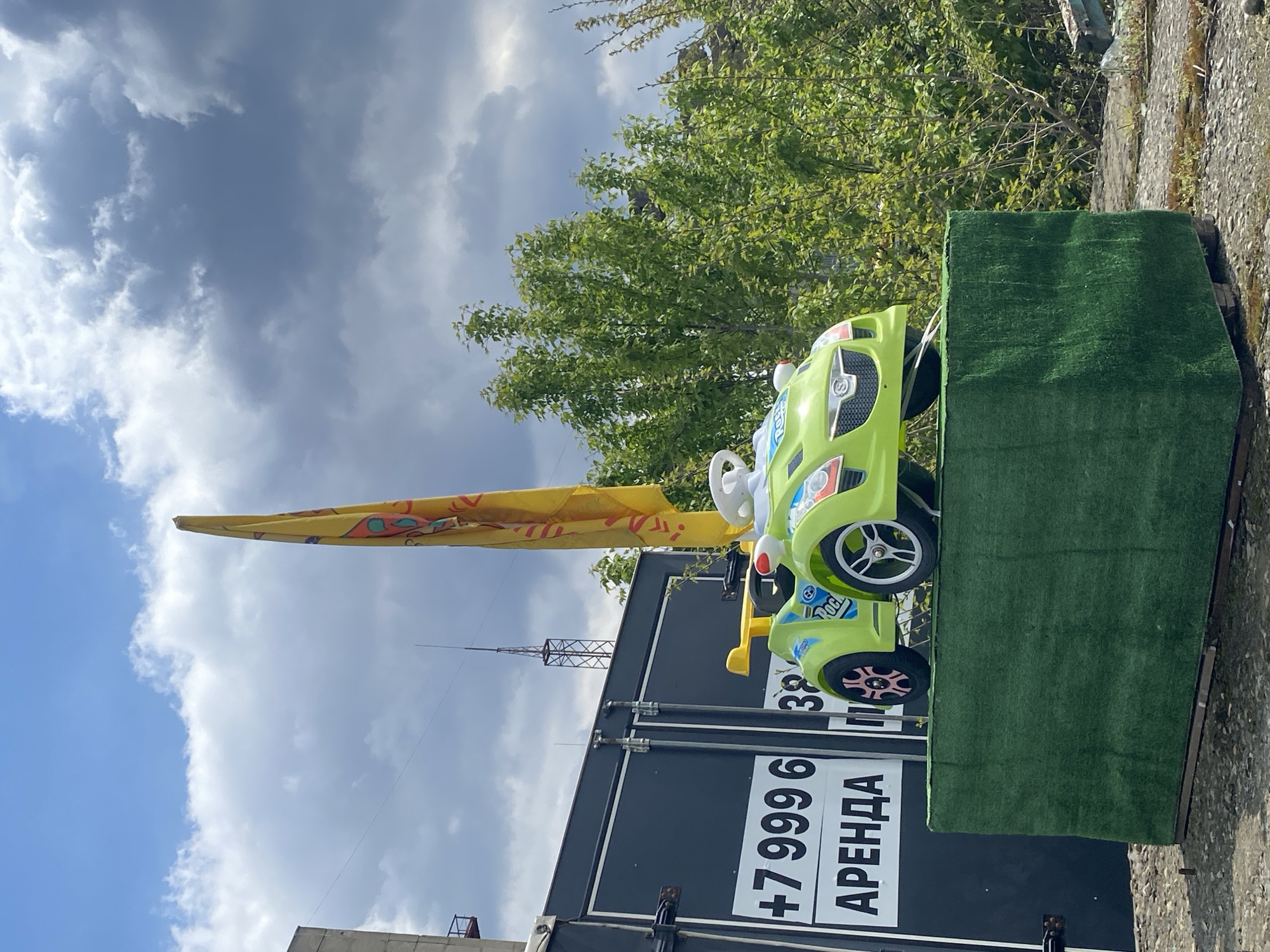 A sideways medium shot shows a green child's toy car with a white steering wheel on top of a short green grassy platform next to a black sign reading "+7 999 6 АРЕНДА". Behind the sign and the car is a yellow flag, a communications tower, some trees, and a sky with clouds. The location is unknown, though the sign is printed in the Russian language.