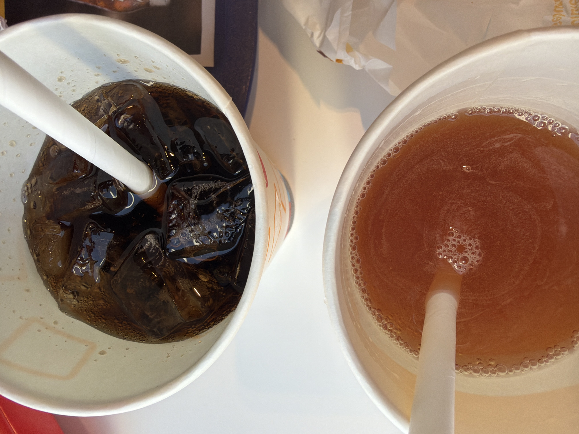 A top-down, close-up view shows two beverages in white paper cups on a light-colored surface, characteristic of a fast-food or casual dining setting in Chuchuligovo, Bulgaria.
The cup on the left contains a dark, carbonated drink, likely a cola, with numerous ice cubes and visible bubbles. A white paper straw is submerged diagonally into the beverage. The outer side of this cup has faint, partially visible red and orange patterned markings.
The cup on the right holds an amber-colored liquid, resembling iced tea or juice, also with a white paper straw. Small bubbles encircle the liquid's edge and gather around the straw.
In the background, a dark blue or black object, possibly a menu or tray, is partially visible behind the left cup. Above and between the cups, crumpled white paper, likely a napkin or food wrapping, is present. Faint, unidentifiable text is barely visible on this crumpled paper. The scene is brightly lit, indicating a daytime setting or a well-illuminated indoor environment. No people are visible.