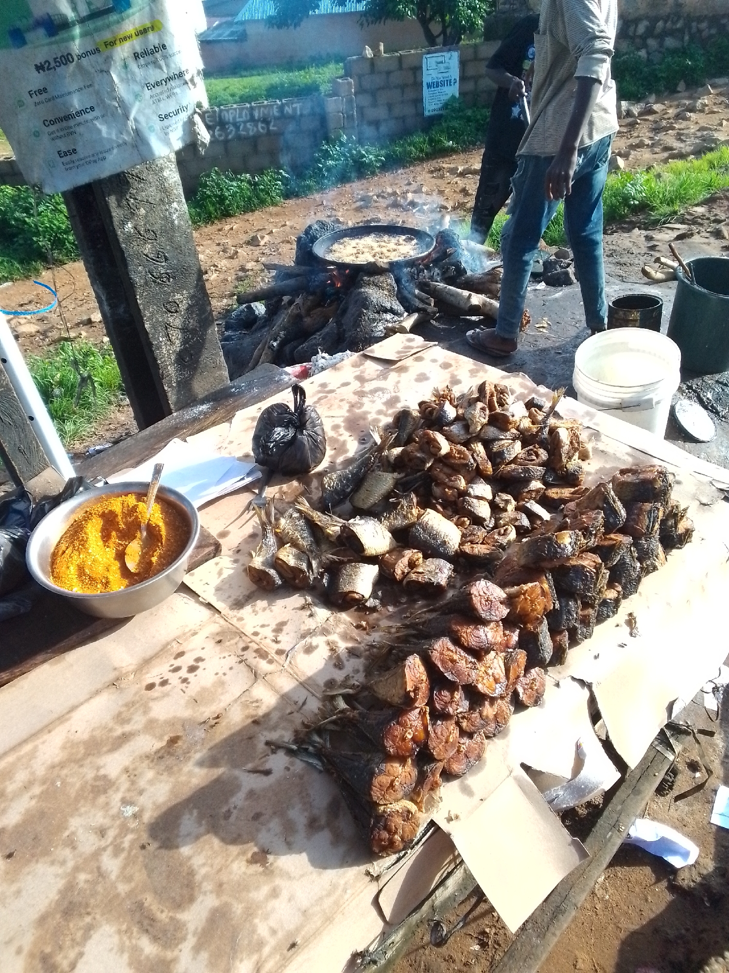An outdoor scene in Jos, Nigeria, depicts a food vendor selling fried fish. The foreground features a table covered with a brown paper cloth, laden with a large pile of fried fish and a metal bowl filled with orange-colored spices. A black plastic bag sits near the bowl, and a spoon rests in the spices. To the right of the fish, two white plastic buckets are visible, one with its lid off. In the background, a man is tending to a cooking fire, with a pan of food over the flames. Another person stands behind him, partially obscured. A post with a weathered sign is on the left side of the frame. The ground is uneven, with dirt and sparse vegetation. The overall impression is of a bustling street food stall.
