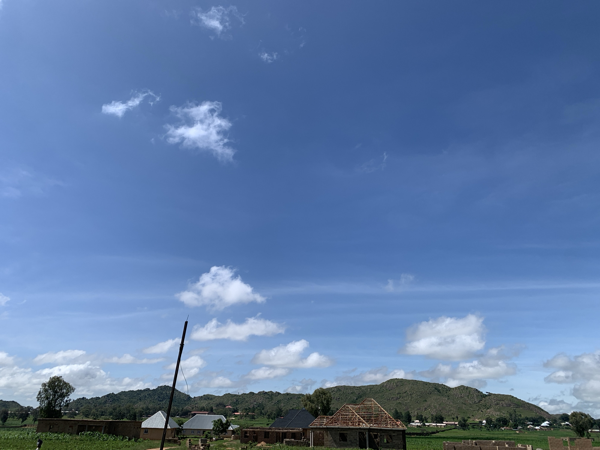 The image captures a rural landscape in Kuru Karama, Nigeria, under a bright blue sky dotted with scattered white clouds. In the foreground, several buildings are visible, some appearing to be under construction. One building in the center features an exposed wooden roof structure, suggesting ongoing work. Adjacent to it, another building has a finished, dark-colored roof. To the left, a building with a white roof is situated. A tall, dark pole stands prominently in the middle ground, extending vertically into the sky. Green fields and trees populate the scene, with rolling, verdant hills forming the backdrop. The overall impression is one of a developing community nestled within a natural, hilly environment.