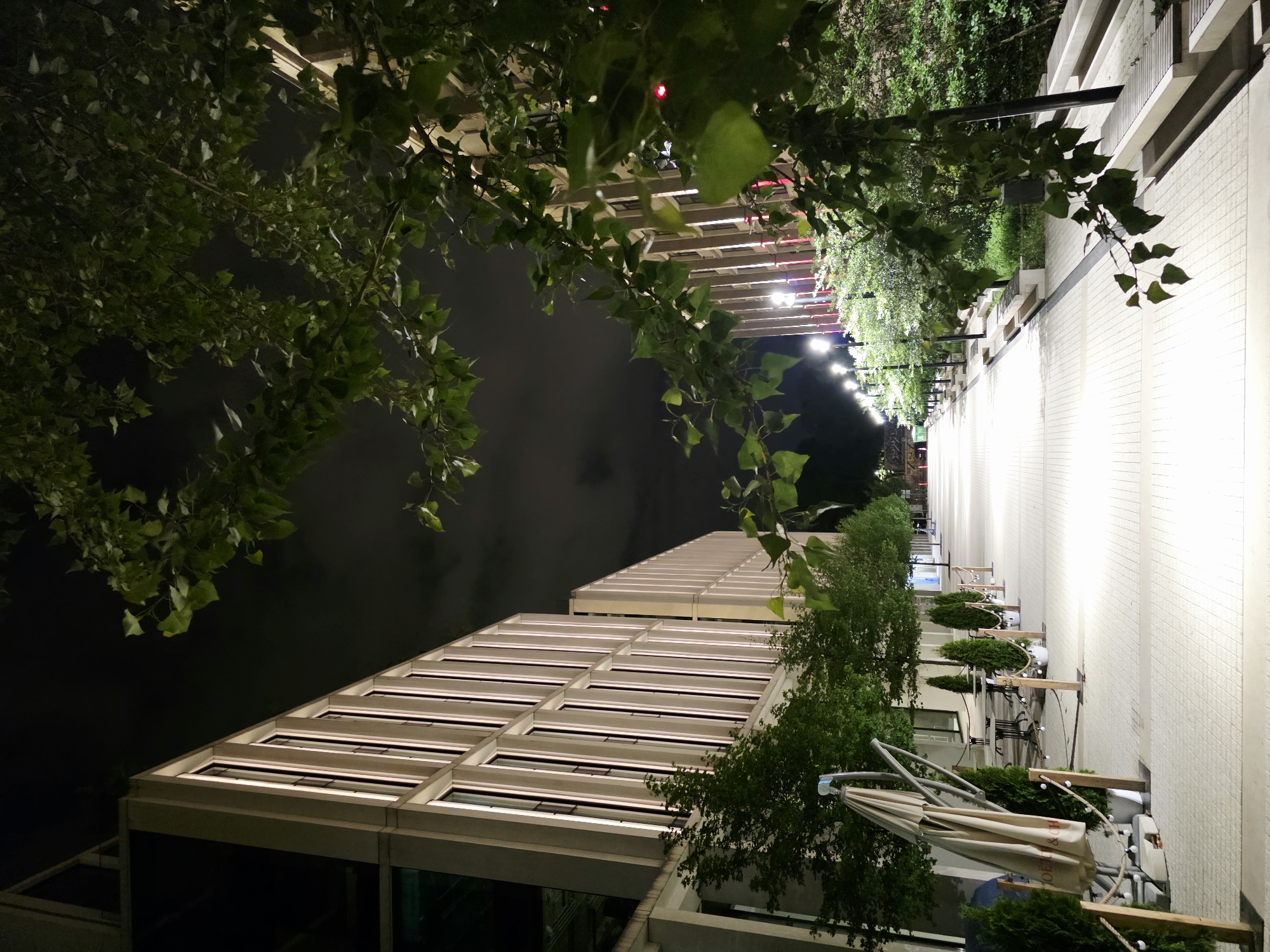 An outdoor perspective at night shows a paved walkway lined with trees and modern buildings. The walkway is illuminated by a series of streetlights casting a bright glow on the ground. On the left, a multi-story building features a facade with vertical elements and numerous windows, some of which are lit. Further down the walkway, another similar building is visible. The right side of the walkway is bordered by lush green trees and benches. In the foreground on the left, an outdoor seating area with tables and chairs is set up, with a large umbrella folded. The sky above is dark and cloudy. The location is the area around the Praga Centrum in Warsaw, Poland.