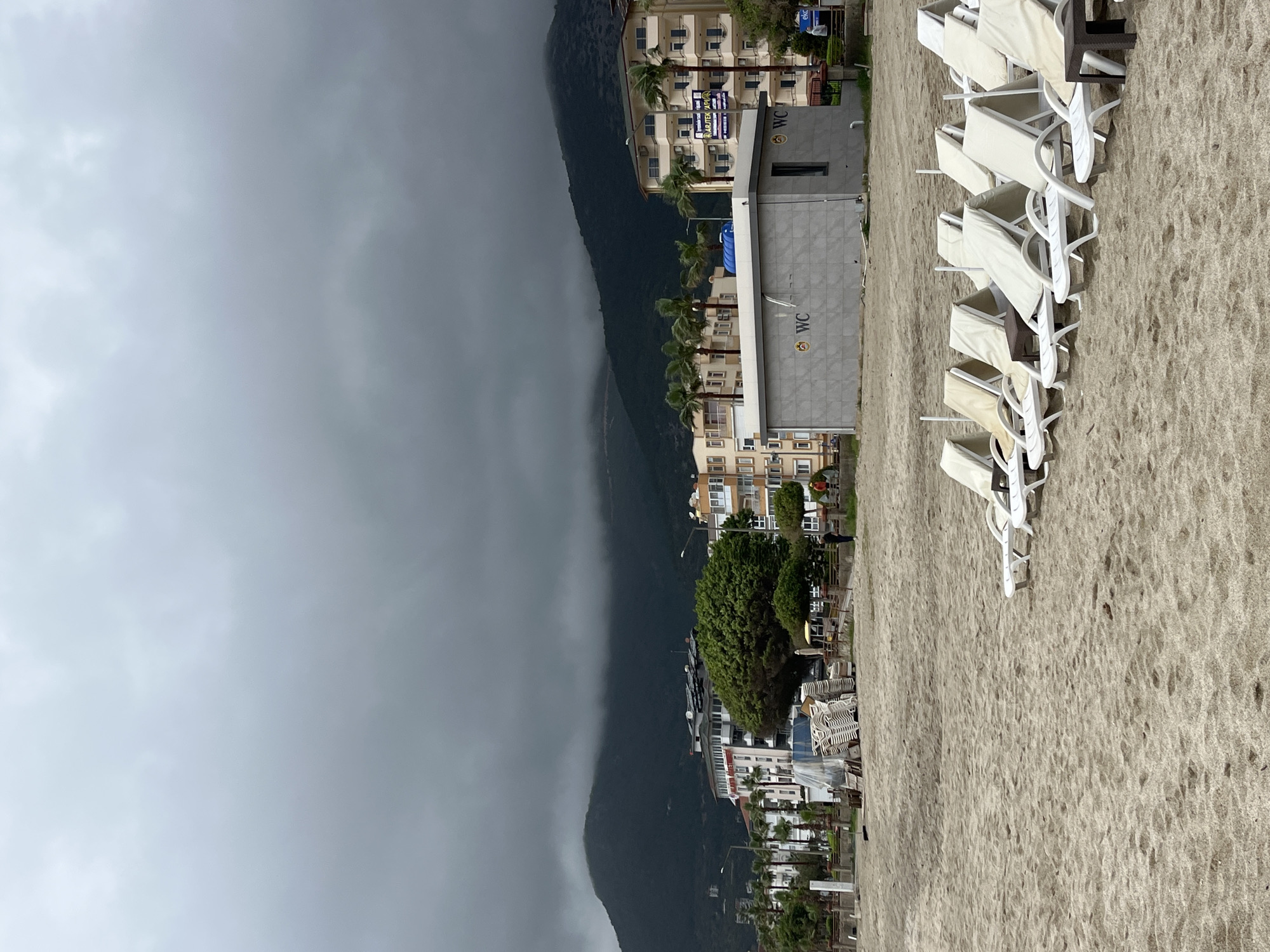 The image captures a beach scene in Alanya, Turkey. Dominating the left side is a large mountain obscured by a heavy, dark cloud cover. Adjacent to the mountain, buildings and palm trees line the coast, suggesting a built-up area. On the right side, the sandy beach is visible, featuring rows of empty white beach chairs. The sand appears to be damp, and the sky suggests a gloomy, possibly rainy, weather condition. A building marked with "WC," an abbreviation for water closet, stands near the beach chairs. The photograph appears to be taken from a slightly elevated position, possibly from a building overlooking the beach. No people are directly visible in the frame.