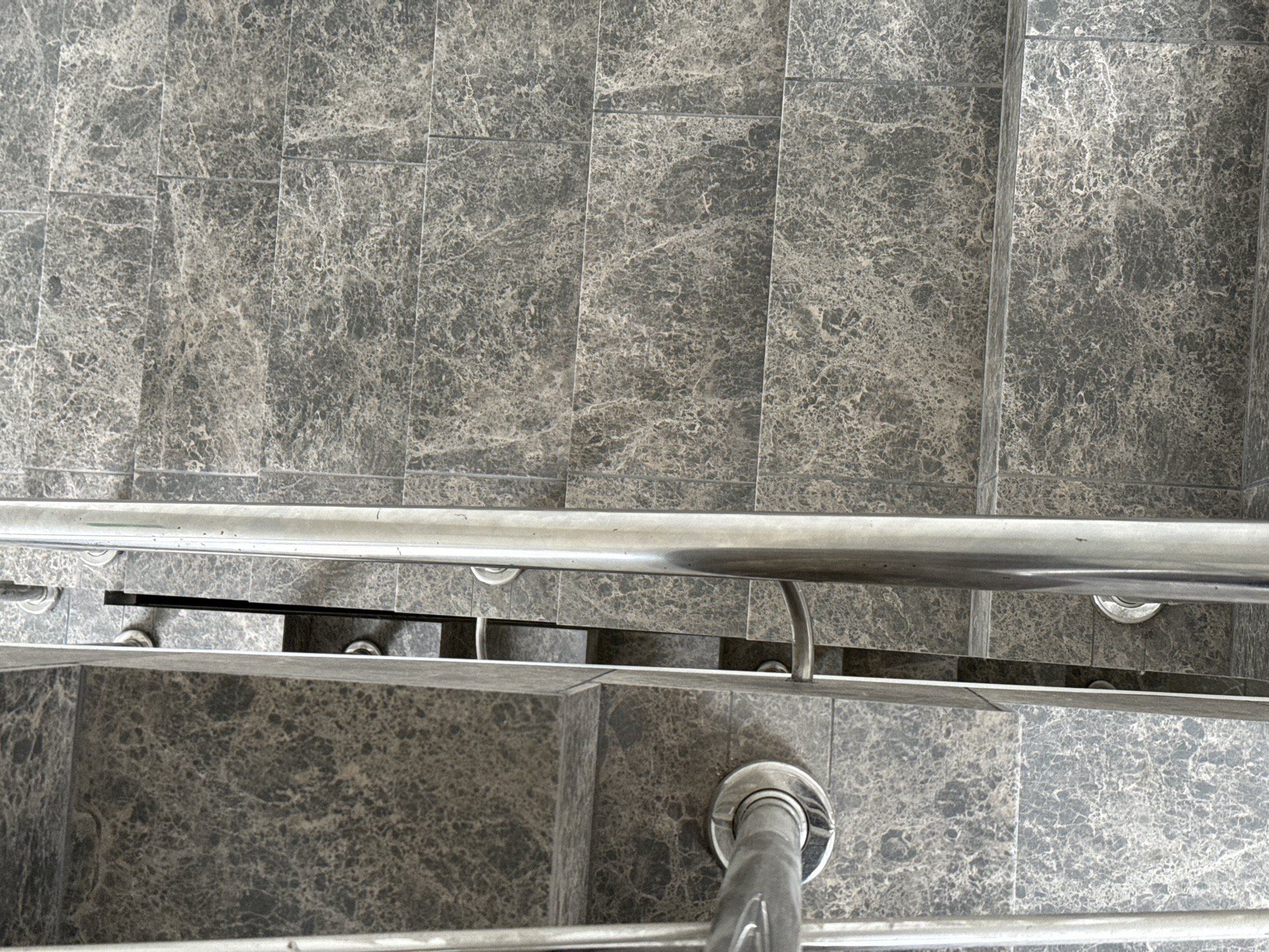 The image shows a close-up, overhead view of a modern, indoor staircase. The steps and risers are covered in dark gray, marbled tiles. A polished metal handrail runs along the right side of the staircase, with a second handrail visible on the left. The handrails are supported by round, metal fixtures attached to the steps. The scene appears clean and well-maintained.