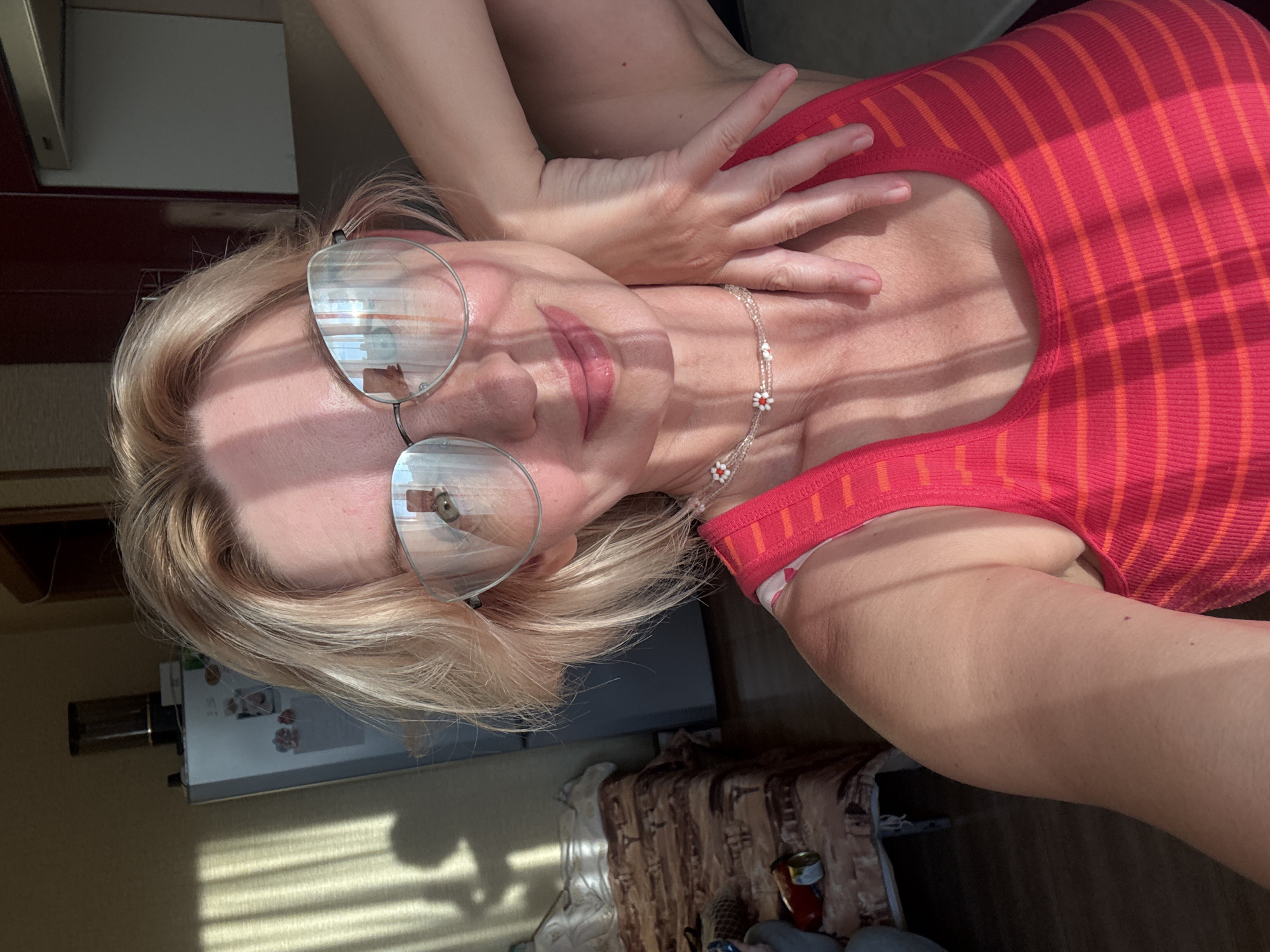 A woman with blonde hair and glasses takes a selfie indoors, possibly in Sochi, Russia, with sunlight casting shadows on her face and upper body. She is wearing a red and orange striped tank top and a delicate beaded necklace with small white and red flower charms. Her right arm is raised and resting on her neck. In the background, a refrigerator, a bed with a patterned comforter, and kitchen cabinets are visible, suggesting a residential setting. The overall lighting is bright, with strong contrasts due to the sunlight.