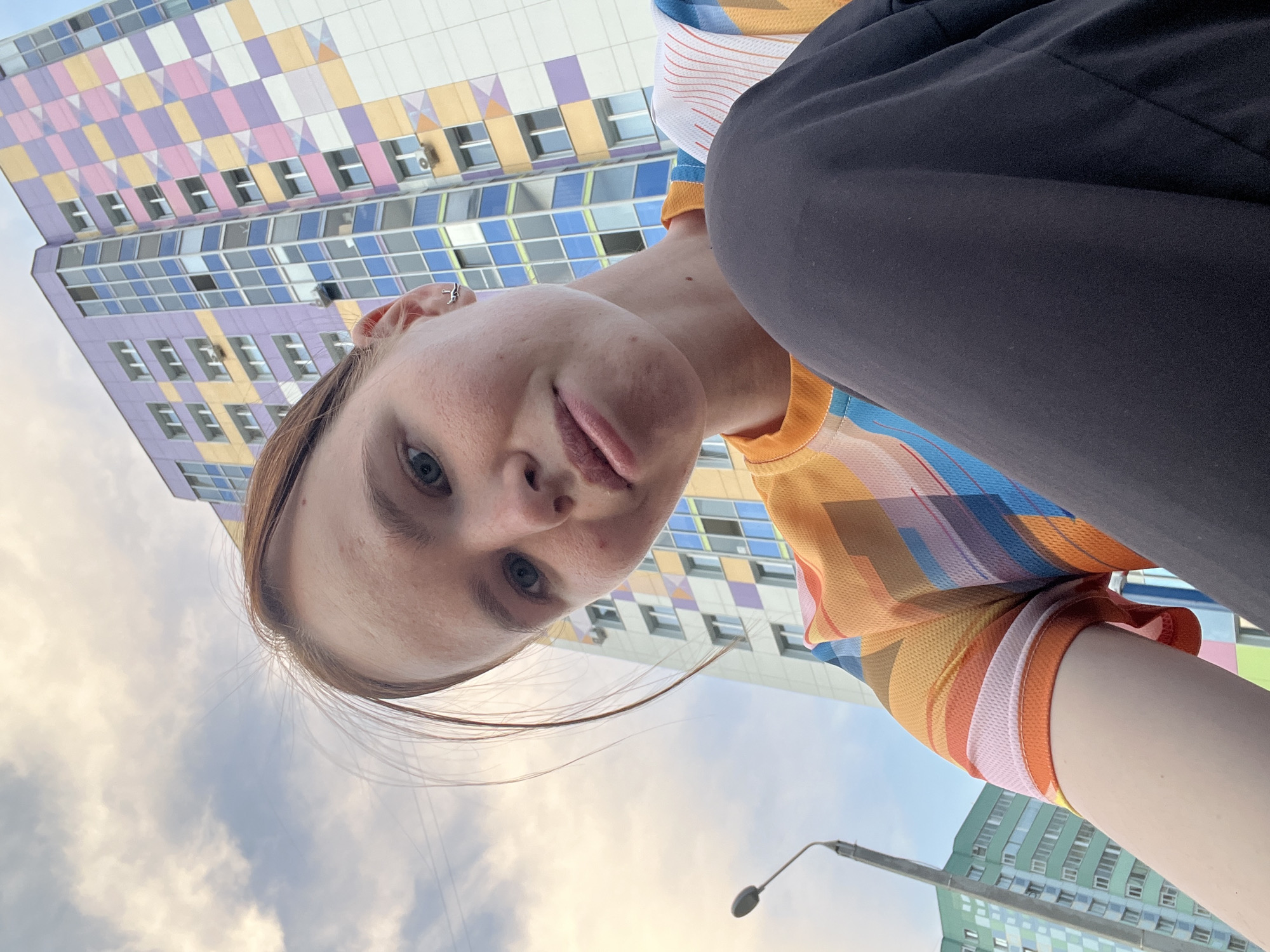 A person is facing downwards toward the camera, which is angled upwards toward them. The person is wearing a multicolored short-sleeve shirt and dark pants. In the background is a colorful, modern building in Perm, Russia, featuring a geometric pattern of purple, yellow, green, and blue. Some windows are visible on the building. The sky is visible with some clouds. A street lamp can be seen partially at the bottom.