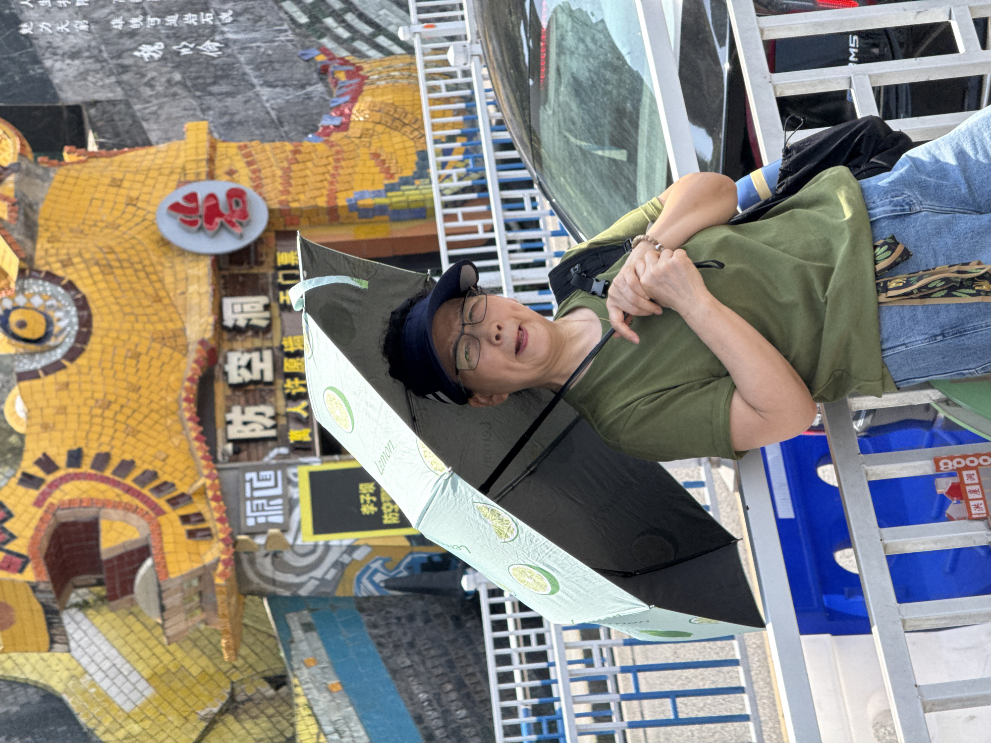 This outdoor image, taken in Yuzhong District, China, features an individual holding an umbrella against a backdrop of a colorful mosaic structure. The subject, an East Asian person appearing middle-aged or older, wears a dark blue cap, glasses, a green short-sleeved shirt, and blue jeans. They are in a leaning or resting position, with hands clasped, and a beaded bracelet on one wrist. A black fanny pack is visible across their chest. The light green umbrella, patterned with lemons and bearing the word "Lemon," is open, providing shade or cover.
The background is dominated by an intricate, yellow-tiled mosaic structure, which features red and blue accents, resembling a fantastical architectural element or sculpture. A circular sign with stylized red characters, possibly "4R," is affixed to this structure. Behind the person, a metallic grate or railing and part of what appears to be a black vehicle or equipment with "MS" and "8000" visible can be seen.
Various Chinese texts are scattered across the scene. On the mosaic structure, prominent phrases include "源防空洞" (Yuan Air-Raid Shelter), "萬人许愿墙无门票" (Ten Thousand People's Wishing Wall, No Ticket), and "防空" (Air Defense). Other visible texts read "魅力无窮 雄魄可追岩石魂" (Endless charm, heroic spirit pursuing the soul of rocks) and "魏心倫" (Wei Xinlun), as well as "李子聪." The scene suggests a public space, possibly a cultural or tourist attraction, on a bright day.