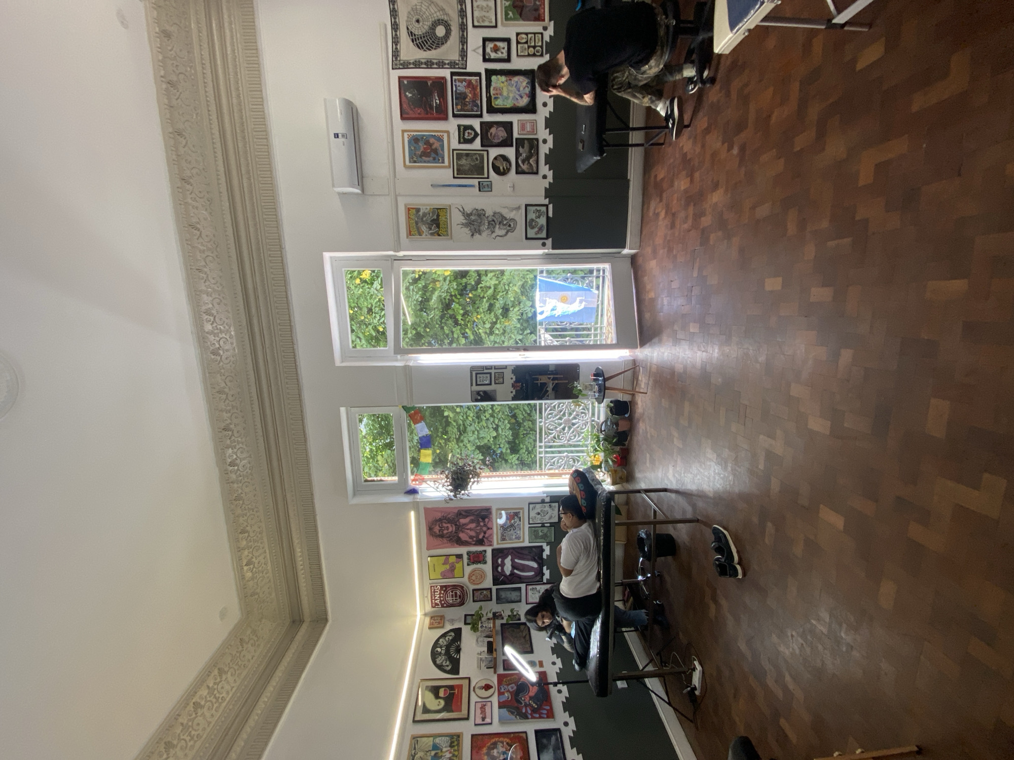 The image depicts the interior of a tattoo studio. There are two tattoo artists working on clients. The studio is decorated with numerous framed pictures and art prints on the walls. The floor is wooden and herringbone-patterned. Natural light enters through a set of open French doors leading to a balcony with a view of trees. An Argentinian flag is visible on the balcony. Based on the style of the architecture and the presence of the Argentinian flag, it is likely that the location is in Buenos Aires, Argentina.