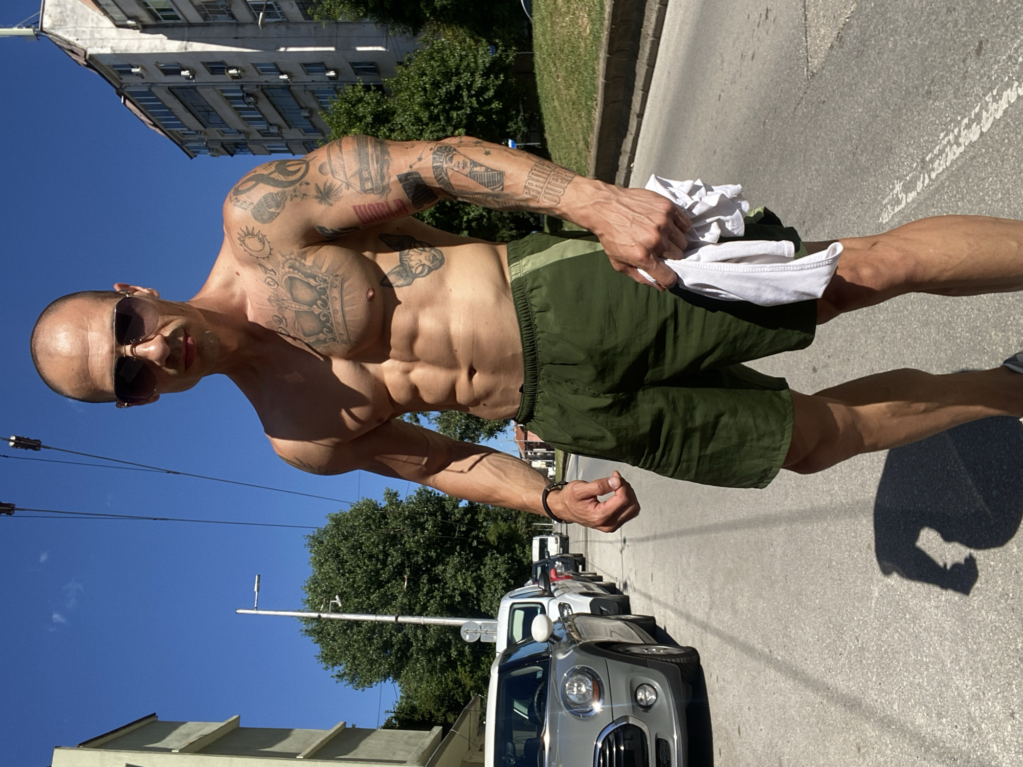 A muscular, shirtless man wearing sunglasses and green shorts stands on a street in Pleven, Bulgaria. He holds a white t-shirt in his right hand. The man has numerous tattoos on his chest and arms. Cars are parked along the street, and buildings and trees are visible in the background. The sky is clear and blue.