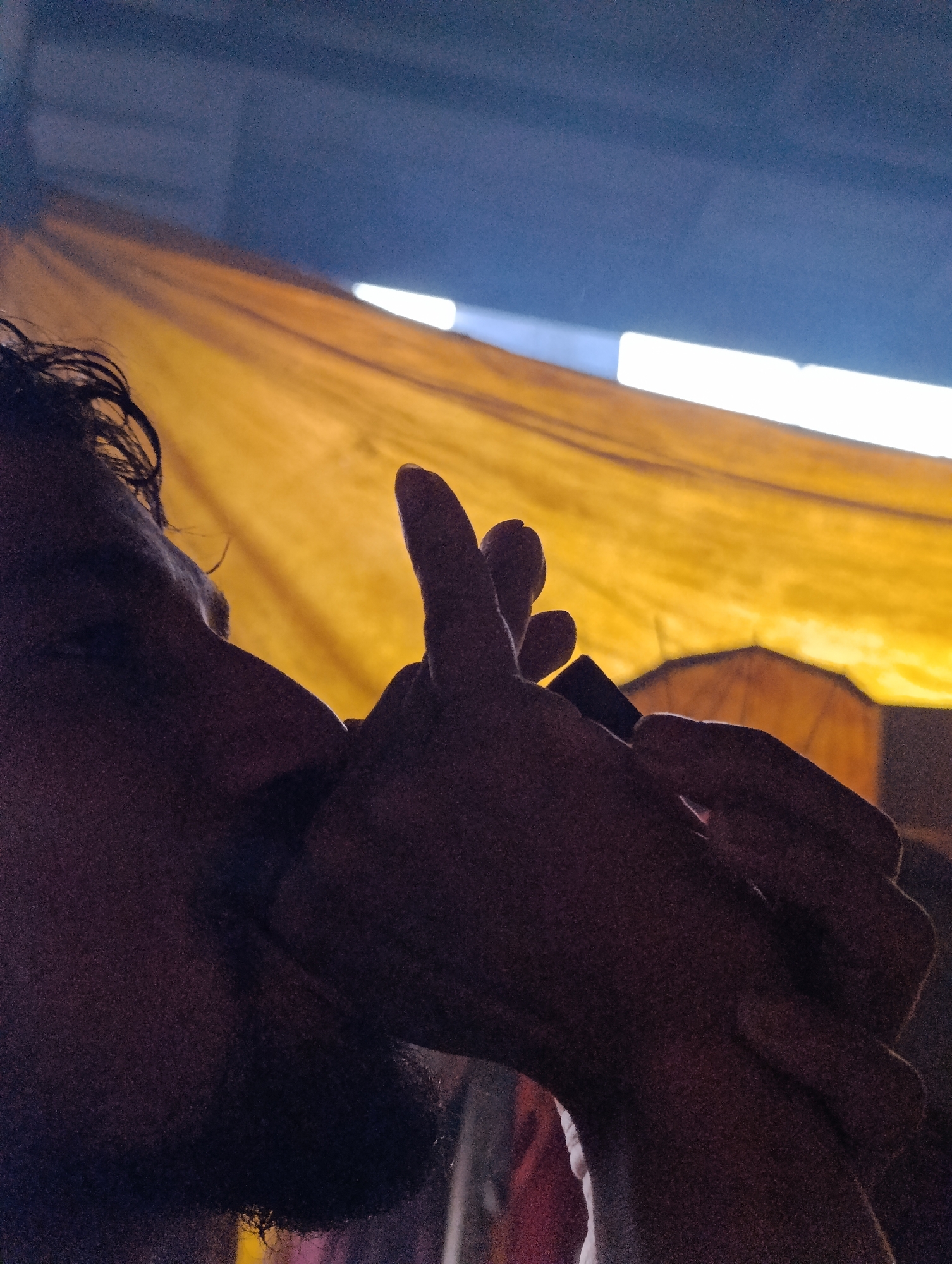 In Vashisht, India, a person, silhouetted against a bright yellow fabric background, is captured in profile. Their face is mostly obscured by shadow, with only a hint of their nose and mouth visible. Their hands are clasped in front of their face, and they appear to be holding a small, dark object, possibly a pipe or a stick, to their lips. The yellow fabric, which seems to be stretched taut, forms a backdrop to the scene, with streaks of bright light piercing through it from above. The lighting suggests an indoor or covered outdoor setting, with the light source likely being artificial or a strong beam of sunlight. The overall mood is one of quiet contemplation or ritual.