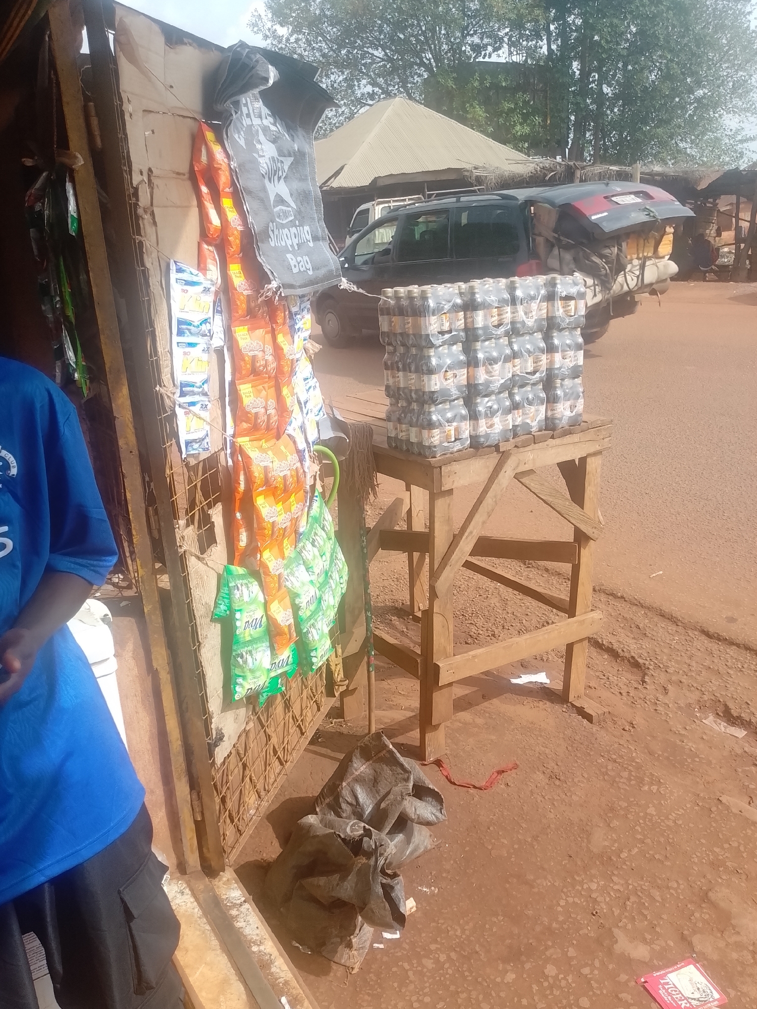 A street scene in Ejule, Nigeria, shows a small roadside shop. A partially visible person in a blue shirt stands on the left. The shop's metal frame displays snacks and a shopping bag. A wooden stand holds stacks of bottled drinks. Debris is on the ground. A black vehicle is in the background on the street. Other structures are visible in the distance. The ground is reddish-brown.
