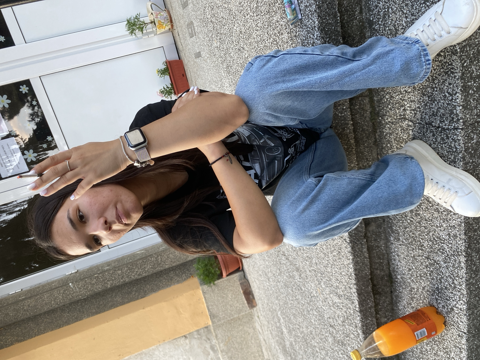 The image captures a young woman, possibly in Hirevo, Bulgaria, seated on concrete steps outdoors. She is wearing a black t-shirt and light-wash denim jeans. Her right hand is raised, holding a cigarette between her index and middle fingers. She is also wearing a silver ring on her right ring finger and a silver smartwatch on her left wrist. A metallic colored charm bracelet with a heart-shaped charm is visible on her left wrist. Next to her on the steps, to her left, is a partially full plastic bottle of orange-colored soda. In the background, a building entrance with glass doors and some decorative plants in pots is visible. The overall lighting suggests it is daytime, possibly late afternoon or early evening, given the shadows.