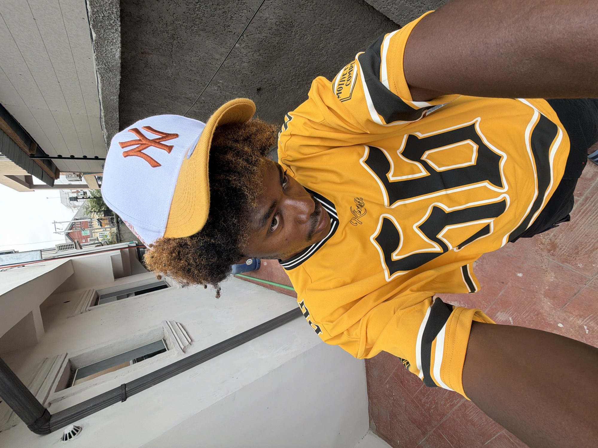 A young man with curly hair is taking a selfie outdoors. He is wearing a white and gold baseball cap with the New York Yankees logo and a yellow sports jersey with black and white trim and large black numbers. The photo is taken from a low angle, showing a white building with window frames and a drainpipe, plus part of a red tiled surface. In the background are buildings in the city of Igboefon, Nigeria.