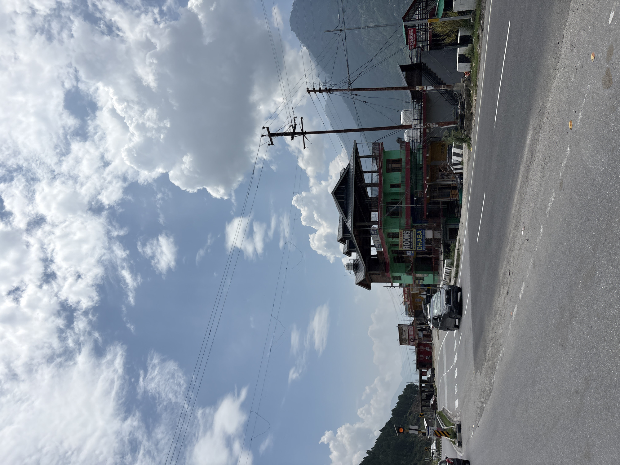 The image depicts a highway scene in Pirdi, India, under a partially cloudy sky. The road curves to the left in the foreground, with white lane markings visible. Several vehicles are present, including a dark SUV driving on the road and other cars parked or driving in the distance. Along the right side of the road, a multi-story building with a green facade and balconies is visible, featuring signs that read "ROOMS" and "DHABA". Another building with "HOME STAY" signage is also present. Telephone poles with extensive wiring are prominent fixtures in the scene. Lush green hills and mountains form the backdrop, with some trees visible on the left side of the image. A traffic light is also positioned near the left edge of the frame. The overall impression is of a busy roadside area in a mountainous region.