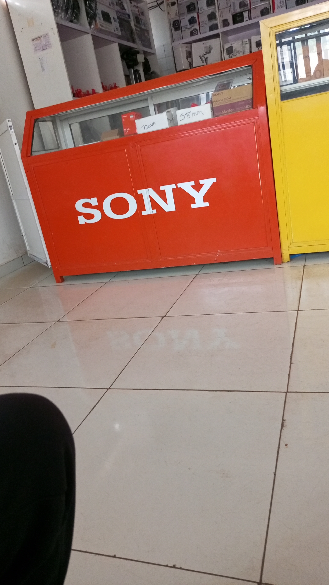 The image captures an indoor scene, likely within an electronics or camera store in Sardauna, Nigeria. The central focus is a vibrant red display cabinet or counter with the word "SONY" prominently displayed in large white capital letters across its front.
Inside the glass top of the red cabinet, several items are visible. These include small boxes, some labeled with lens sizes such as "72mm" and "58mm," suggesting camera filters or accessories. Other boxes inside bear the "SONY" brand name, with additional text like "PHDVM-63RM," "DigitalMaster," and "HDV," indicating video recording media or related products.
Behind the red cabinet, white shelves are filled with numerous stacked cardboard boxes. These boxes appear to contain electronic goods, possibly cameras, lenses, or other photography and video equipment, with some displaying brand logos such as Sony and faintly, Nikon, suggesting a multi-brand retail environment.
To the immediate right of the red cabinet, part of a yellow cabinet or display unit is visible. The floor is covered with light-colored, possibly off-white, ceramic tiles, which reflect the ambient light, indicating a clean and well-lit space. In the bottom left corner, a dark, blurred shape, possibly a person's leg or clothing, is partially visible, suggesting the perspective of someone standing or sitting near the display.
The overall impression is that of a professional retail setting dedicated to electronics, particularly photographic and video equipment. No specific activity is taking place, and the time of day is not indicated by natural light, but rather by indoor lighting.