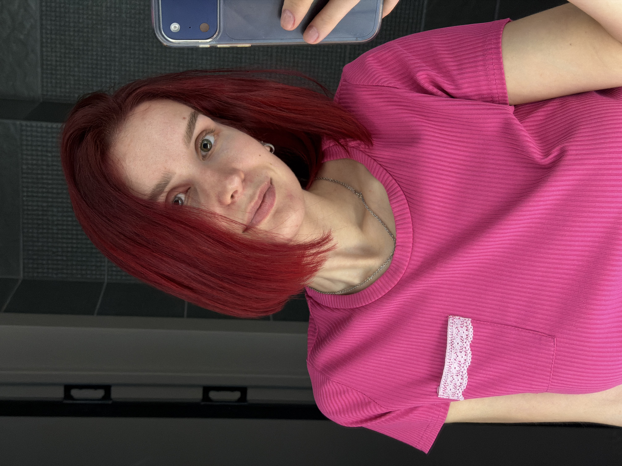 A woman with red hair wearing a pink, ribbed, short-sleeved shirt with a small lace-trimmed pocket is taking a selfie indoors. The background appears to be a tiled wall over what looks like a baseboard heater. The location is specified as Chaikovsky, Russia.
