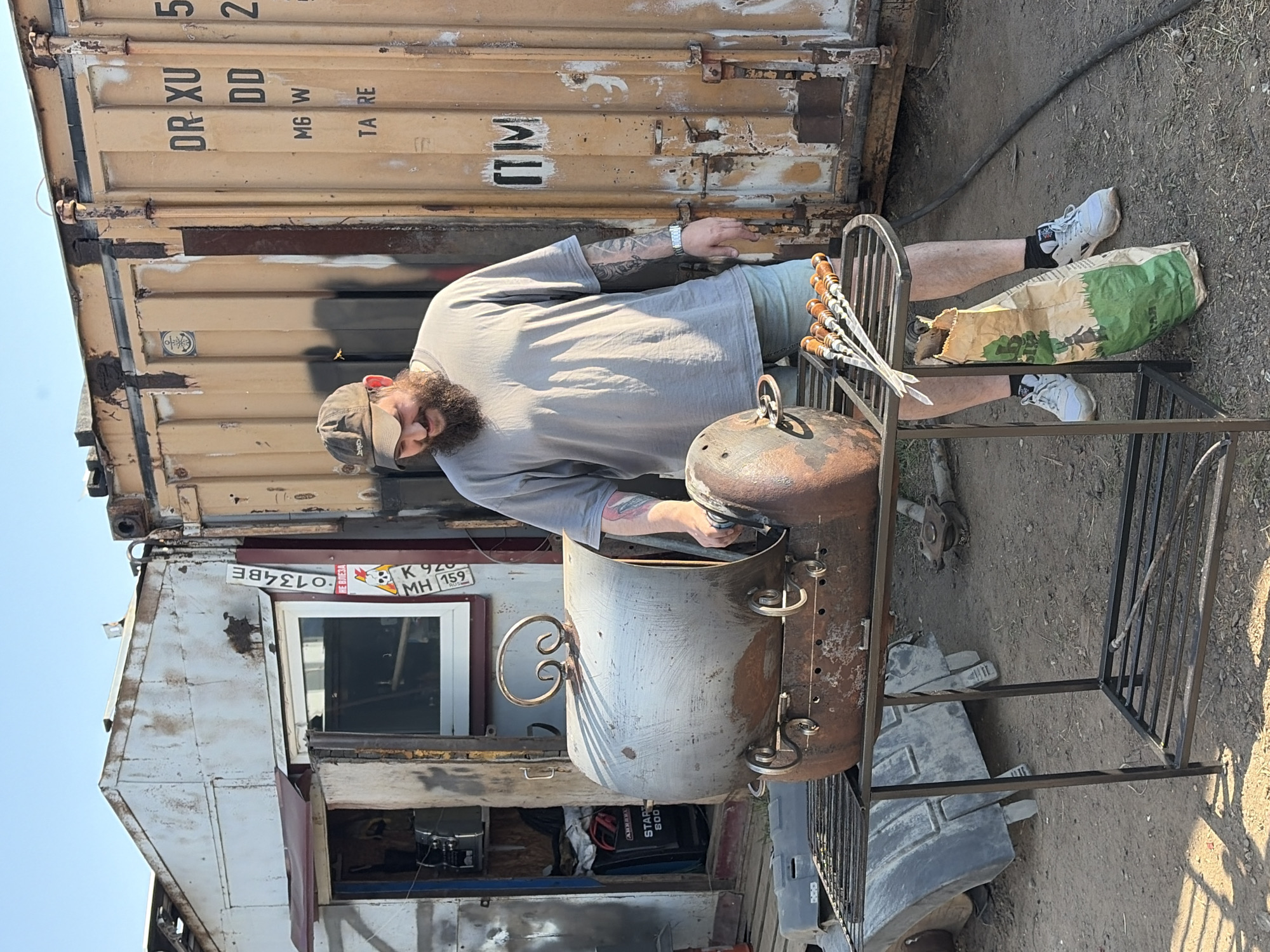 The image shows a man barbecuing next to a container in what appears to be an outdoor setting. The man, wearing a gray t-shirt and a hat, is tending to a cylindrical metal barbecue grill. Skewers are placed on the grill stand next to him. A bag of charcoal is also visible. Behind him, there's a shipping container and what seems to be a modified structure with a window and license plate that reads "K 920 MH 159" and "O134BE." Based on the license plate, which is a Russian format, the location can be identified as Chaikovsky, Russia. There are other metal objects and parts scattered around the ground, adding to the general outdoor environment. The weather appears to be sunny.