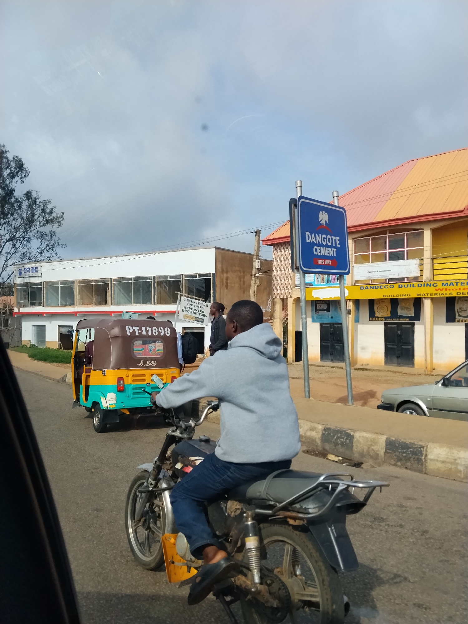 In Gura Top, Nigeria, a man is seen riding a motorcycle down a paved road. He is wearing a grey hooded sweatshirt and blue jeans, with black sandals. In front of him, a yellow and teal auto-rickshaw with a brown canopy is traveling in the same direction. The auto-rickshaw has the license plate "PT-17898" and the words "D. Boy" written on its side.
To the right of the road, there is a building with a bright yellow and orange roof. A blue sign with an eagle emblem and white text reading "DANGOTE CEMENT THIS WAY" is prominently displayed on a metal pole in front of the building. Another building with a white facade and large windows is visible behind the auto-rickshaw, with signage that includes Korean characters ("한국마트" - Hanguk Mart) and English text. Other storefronts with various advertisements are also present along the street. A silver car is parked on the right side of the road in the background. The sky is cloudy.
