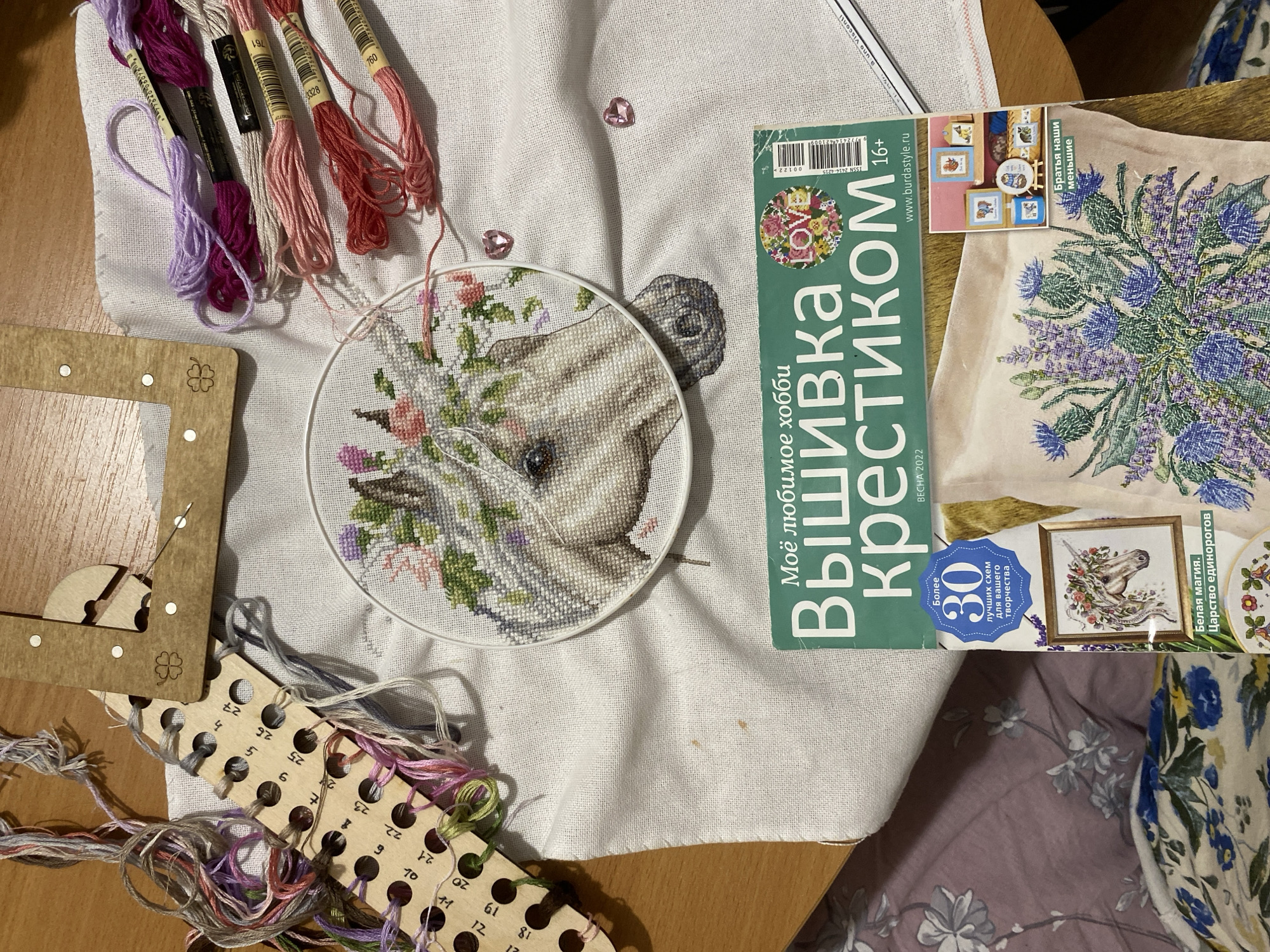 This image shows a close-up of a cross-stitch project in progress. The cross-stitch pattern features a horse with flowers around its neck and head, held in an embroidery hoop. To the left of the hoop is a wooden thread organizer, along with a holder for the thread with a needle. Beside the hoop are spools of embroidery thread in shades of pink, purple, and beige. On the upper part of the image are two pink, heart-shaped decorative objects. To the right of the embroidery is a cross-stitch magazine, with the headline in Russian, containing a cover image of a similar cross-stitch. Under the magazine, a printed fabric with a lavender floral pattern is partially visible. The scene is lit by natural light and the table beneath is visible.