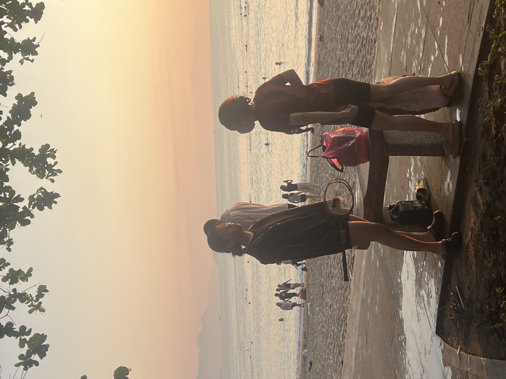 The image depicts a serene beach scene during either sunrise or sunset, characterized by warm, hazy light. In the foreground, two individuals are standing on a paved walkway adjacent to a sandy beach. One person, wearing a dark shirt and shorts, holds a badminton racket and looks towards the sea. Slightly behind them, another person in a red shirt and shorts also faces the water. A red bag rests on a low wall or bench between the two. Various shoes are visible on the ground nearby, including a pair of dark sneakers and some lighter footwear.
The paved area gives way to a sandy beach where several other people are visible, some standing by the shoreline and others wading or swimming in the calm sea. Distant mountains are faintly silhouetted against the horizon under the golden sky. The top left corner of the frame is adorned with dark green foliage from an overhead tree, with a small bird barely discernible in the sky within one of the cropped images. The overall atmosphere suggests a leisurely and peaceful recreational activity by the sea, characteristic of a location like Nha Trang, Vietnam, as provided in the context. There is no visible text in the image.