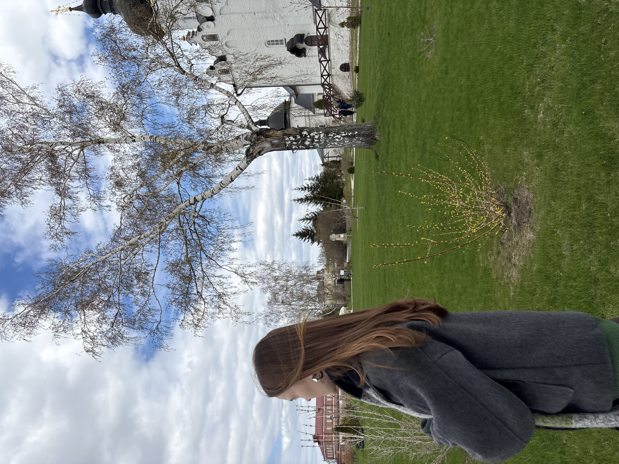 A waist-up view of a woman with long brown hair in a gray coat, turned to her left, with a blurred background. Beyond her are flowering shrubs in a grassy field near a white building with dark domes in the historical village of Sviyazhsk, Russia. A birch tree with bare branches stands between the woman and the building. The sky is partially cloudy.