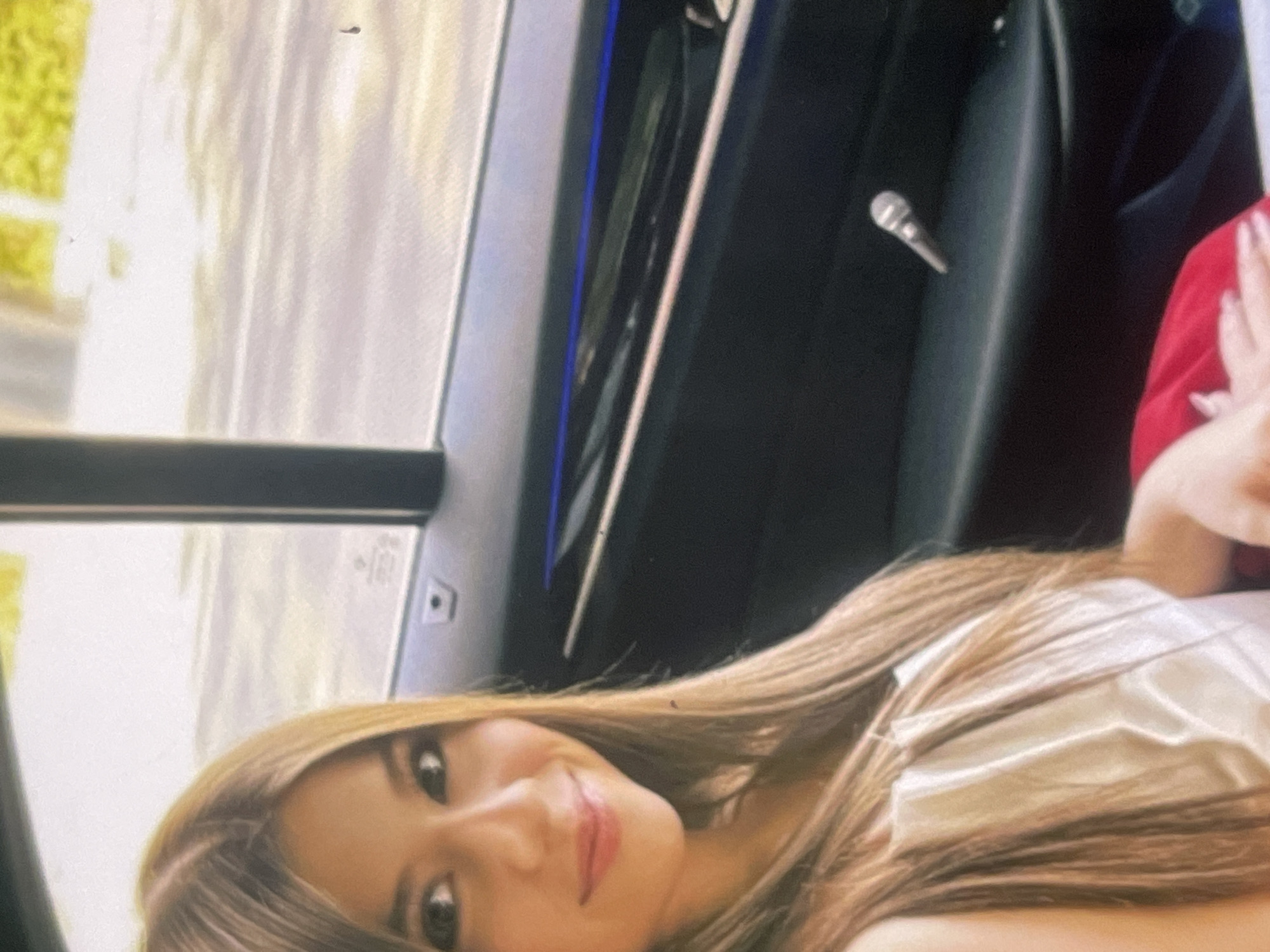 A young woman with long, light brown hair is pictured in what appears to be the interior of a vehicle, possibly a car, in Poltava, Ukraine. She is smiling directly at the camera, conveying a cheerful and relaxed mood. Her head is positioned towards the left side of the frame, and she is wearing a light-colored top with a visible sheen, suggesting material like satin or silk.
Through the car window on the left, a brightly lit, blurred outdoor scene is visible, dominated by green foliage or trees, indicating it is daytime. The car's interior is dark, with a vertical blue ambient light strip running along the door panel on the right side of the image. A small, metallic, microphone-like object or a specific car feature is attached to this door panel. A portion of another person's arm or hand, wearing a red garment, is partially visible on the far right edge of the frame. On the car window, a faint, small symbol or icon, possibly a car safety mark or manufacturer's detail, can be seen, but no distinct text is legible.