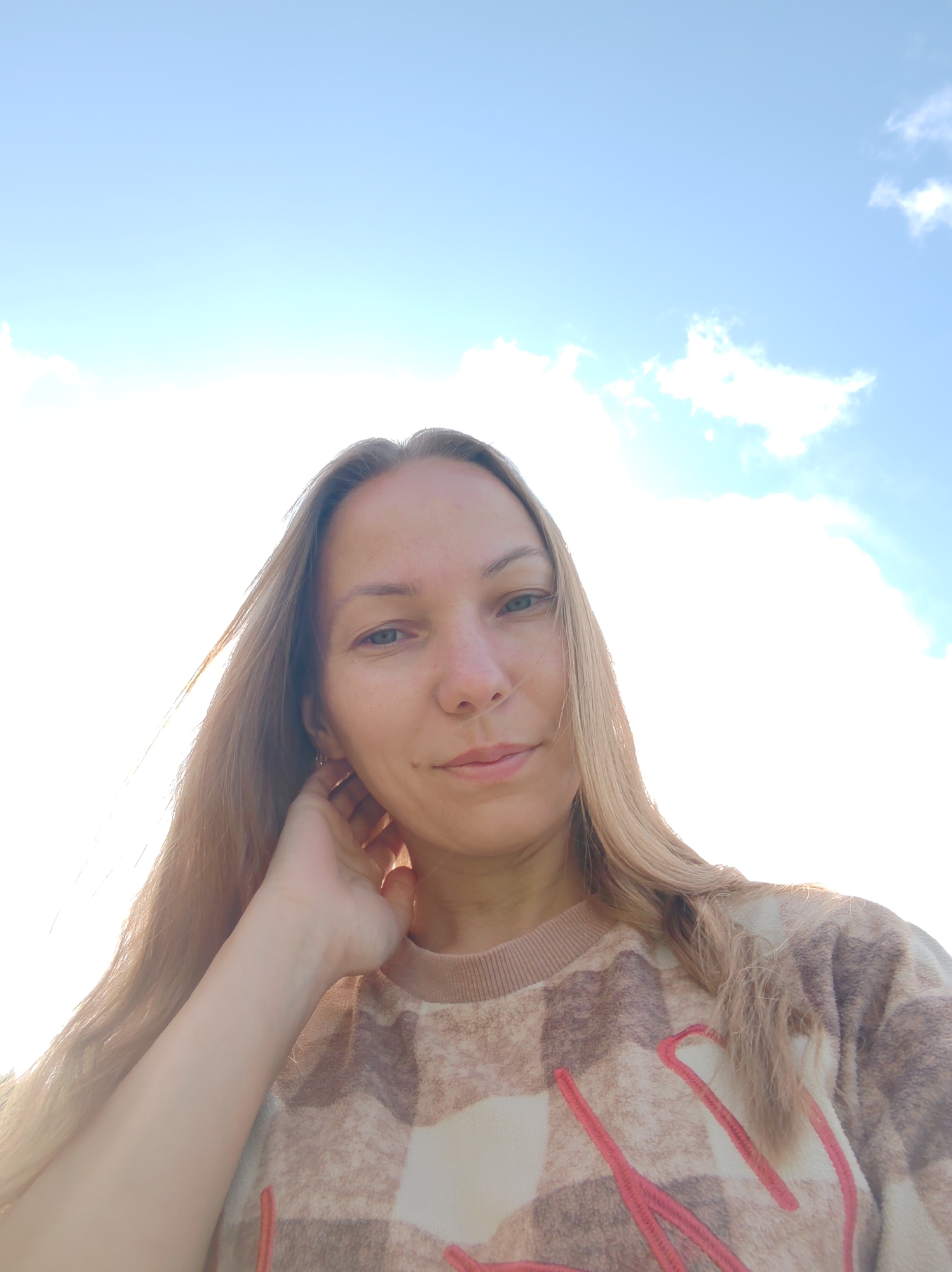 In a low-angle, outdoor selfie, a fair-skinned woman with long, light brown hair and green eyes gazes at the camera with a slight smile. Her left hand is raised to her neck, and she is wearing a cream-colored, plaid-patterned sweatshirt with pink embroidered lettering. The background consists of a bright, hazy sky with a few scattered white clouds. The image is set in Чухлэм, Russia, under an open sky.