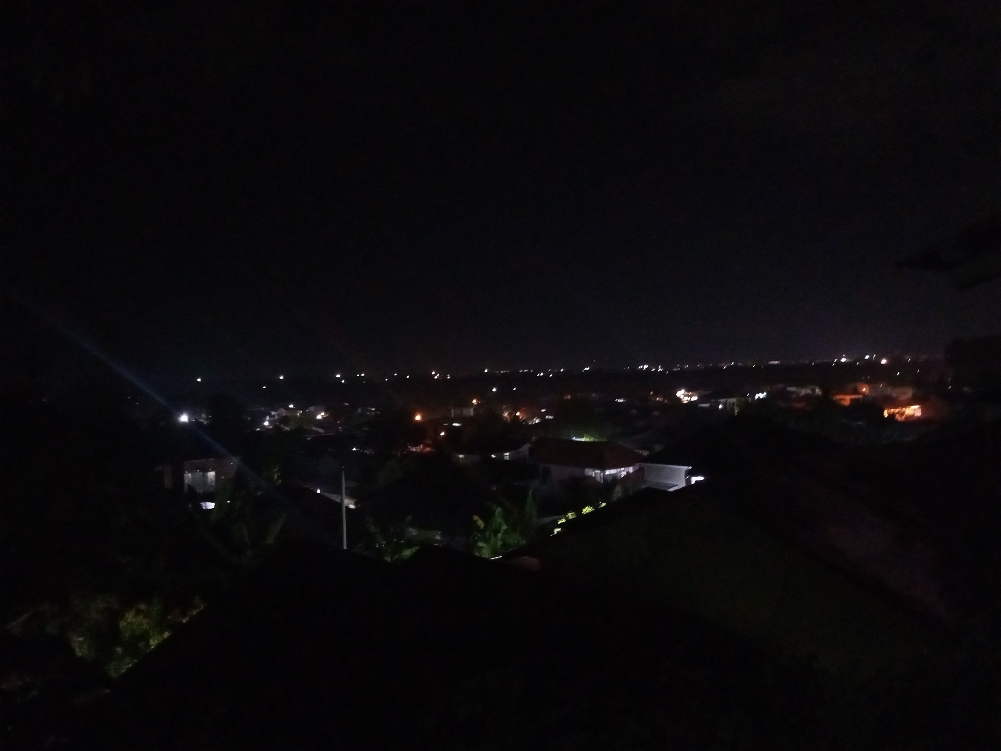 This is a nighttime view of what appears to be a residential area in Lhokseumawe, Indonesia. The scene is dominated by darkness, with clusters of light suggesting houses and streetlights scattered across the landscape. Roofs of houses are visible in the foreground. The city lights stretch along the horizon.