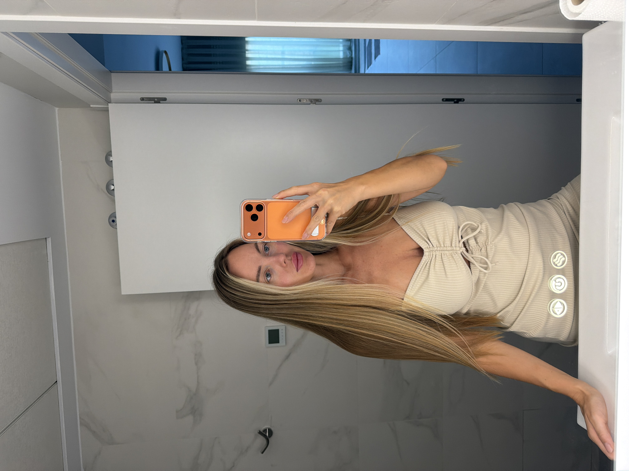 A woman is taking a selfie in a modern bathroom. She is standing in front of a large mirror, which reflects her and the surrounding environment. The woman has long blonde hair and is wearing a light-colored, sleeveless dress with a ruched detail at the bust. She is holding up an orange smartphone to take the picture. The bathroom features marble-patterned walls and a white sink. In the background, a doorway leads to another room with blue curtains and subtle lighting. The location is identified as Finestrat, Spain.