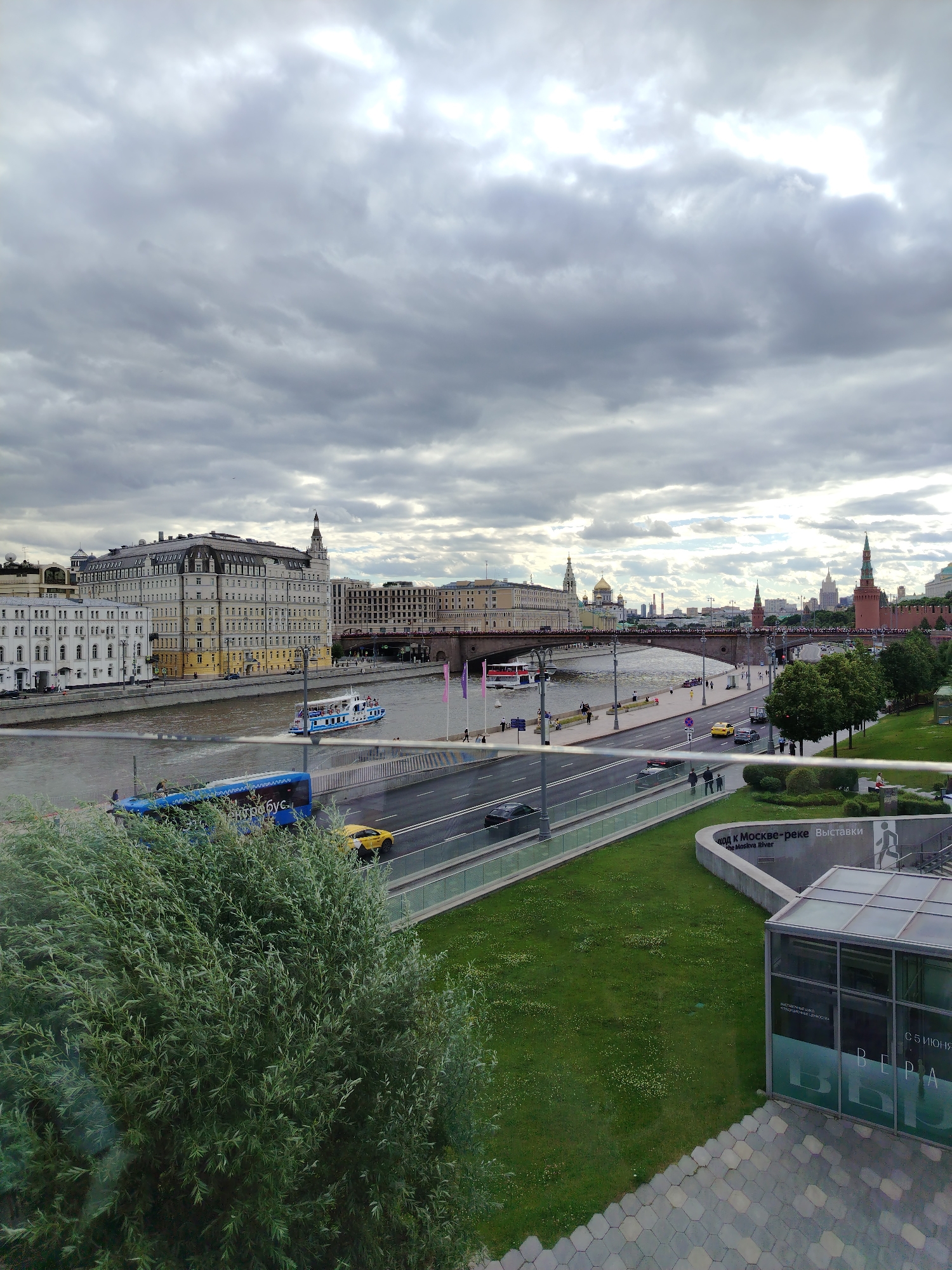 The image depicts a cloudy day in a city, likely Moscow, Russia, given the architecture and the Cyrillic text. A wide river flows through the center of the frame, with a large, multi-story building on the left bank. A white and blue tour boat is on the river, moving from left to right. Across the river, a long bridge spans the water, connecting to a cityscape that includes numerous buildings, some with distinctive onion domes. On the right side of the frame, a modern building with a glass facade is partially visible, situated at the bottom right. In front of this building is a landscaped area with green grass and trees. A road runs parallel to the river, with a blue bus and a yellow taxi driving on it, along with a few pedestrians. The overall impression is a view of a bustling urban waterfront on an overcast day.