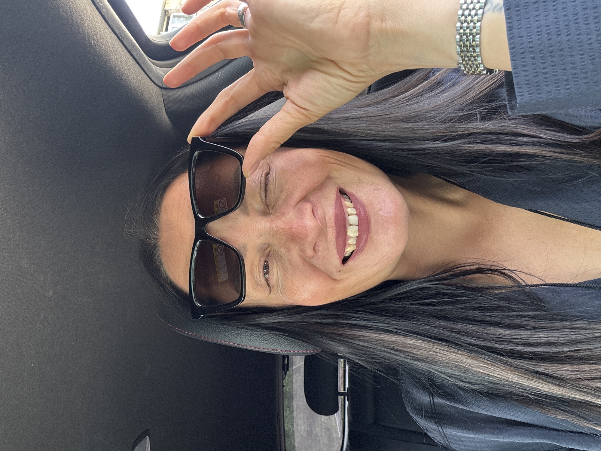 A woman is inside a car, holding sunglasses with one hand in front of her face. She is smiling and has gray and black hair. She is wearing a silver bracelet. The car has a black interior, and the location appears to be outdoors. There is no information to indicate the location is in the city of Chaikovsky, Russia.