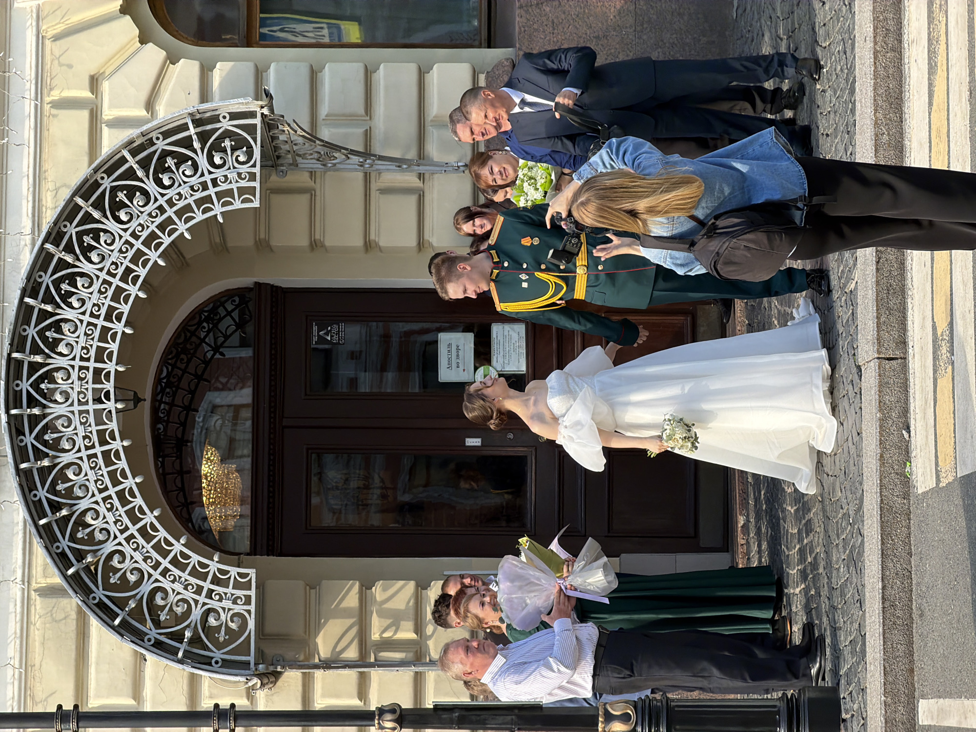 The image depicts a wedding party on a crosswalk in front of an ornate building. A bride in a white gown and a groom in a military uniform are the central figures. Several people, including family and a photographer, surround them. The building's architecture is detailed, with a decorative archway. The location is likely in Saint Petersburg, Russia.