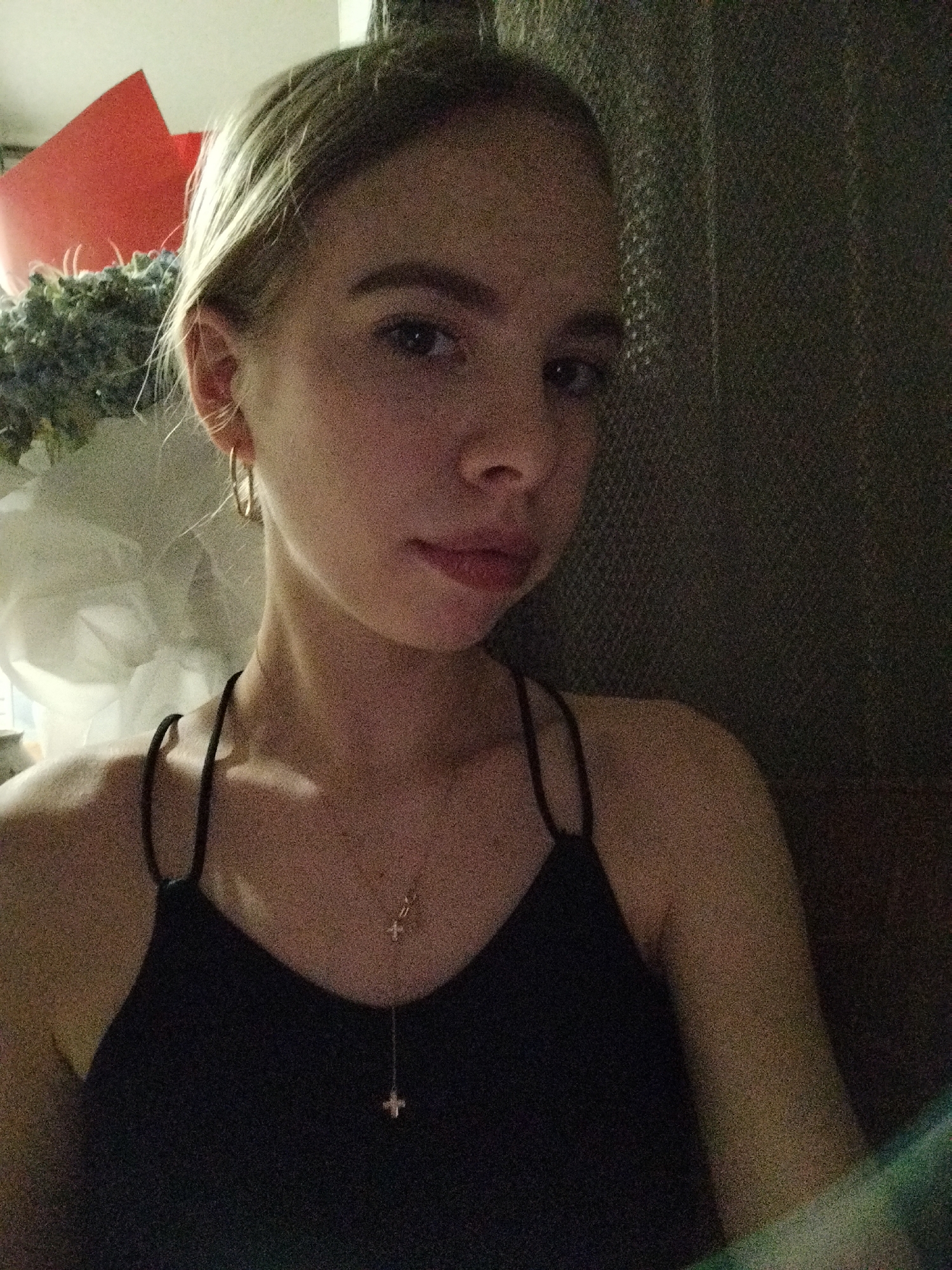 A young woman with blonde hair pulled back is captured in a close-up selfie. She is wearing a black camisole and a delicate necklace with a cross pendant. Her left ear is adorned with a gold hoop earring. The background is dimly lit and textured, suggesting an indoor setting. To the left of the frame, a hint of a red object and some greenery is visible, possibly part of a bouquet or decoration. The overall lighting is soft and warm, with some graininess in the image. The scene appears to be set in an unspecified location within the city of Unknown.