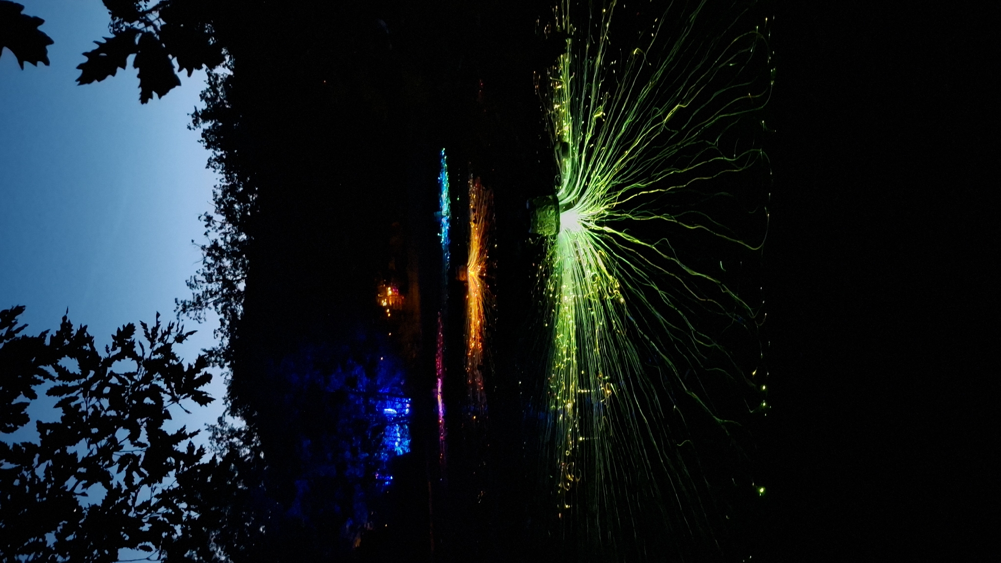 The image depicts a dark scene in Bukovik, Montenegro with trees silhouetted against a blue sky in the upper left corner. To the right of the trees, the scene transitions to near darkness. A series of illuminated objects are visible against the dark background. On the right, a large green light source emanates numerous thin, radiating lines. Moving leftward, there is a vertical arrangement of lights displaying blue, orange, and pink hues. Further to the left, additional blue lights are visible.