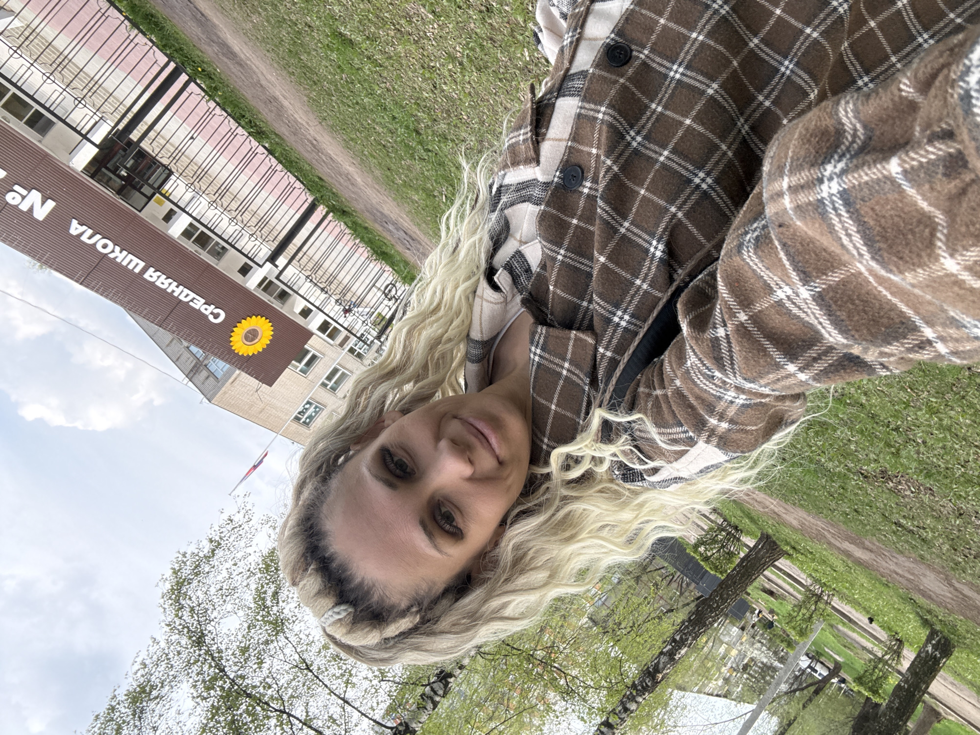 A woman with long blonde hair is posing for a selfie in Заволжье, Russia. She is wearing a brown and white plaid jacket. Behind her is a building labeled "СРЕДНЯЯ ШКОЛА №1" (Secondary School No. 1) with a sunflower emblem. A fence runs along the side of the building. Trees with fresh green leaves are visible in the foreground. A path and grassy area are also visible. The sky is cloudy.