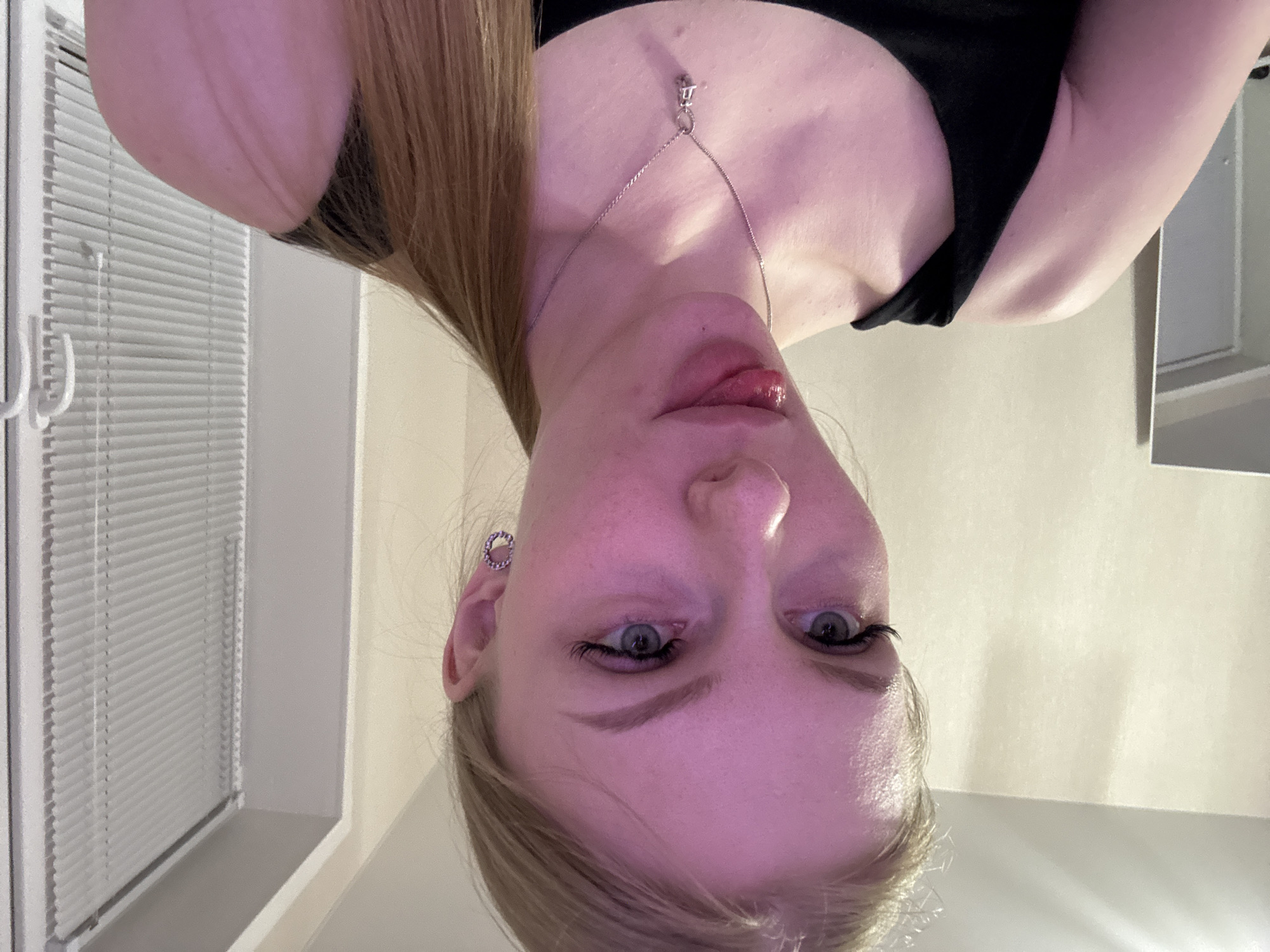 The image shows a woman's face and upper torso, rotated 180 degrees. She has fair skin, blue eyes, and light brown hair. She is wearing a black top and a silver necklace with a pendant. There is an earring visible in her left ear. In the background, there are white blinds covering a window and a light-colored wall. There are no indications that this image was taken in Perm, Russia. The location is indeterminate based on the provided image.