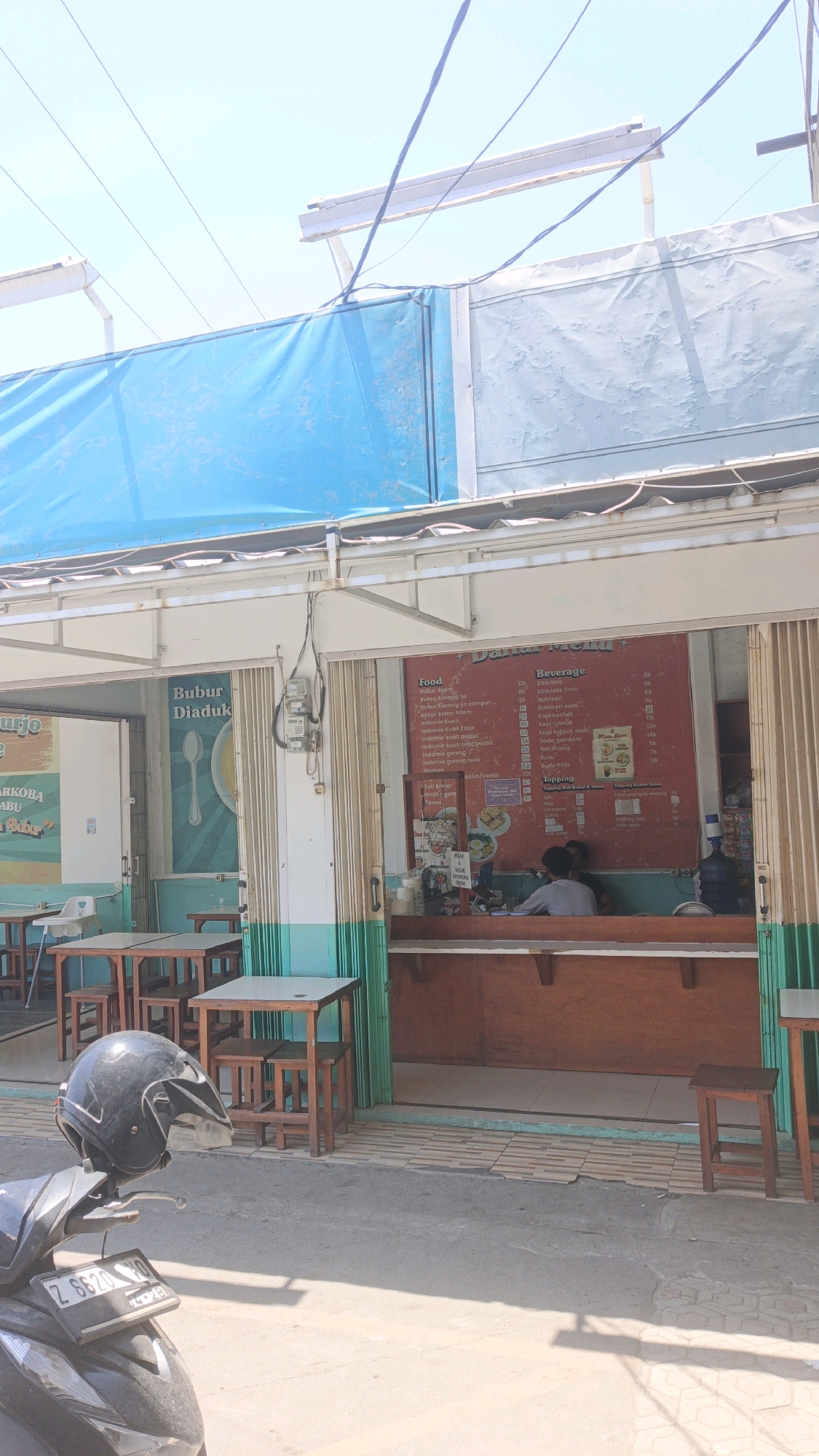 The image depicts the exterior of a small food establishment in Desa Pagandaran, Indonesia, under a bright, clear sky. The building features a blue tarp covering the upper section and a white metal awning. A menu board is prominently displayed on the right side, listing "Food" and "Beverage" options with prices. A wooden counter with seating is visible inside. Two individuals, likely staff or customers, are seated behind the counter. In front of the establishment, several wooden tables and chairs are set up. To the left, a motorcycle helmet rests on the handlebars of a parked motorcycle, whose license plate reads "Z 6620 VO". The ground is paved with tiles and concrete, with shadows cast by the sun. The overall scene suggests a casual, open-air eatery.