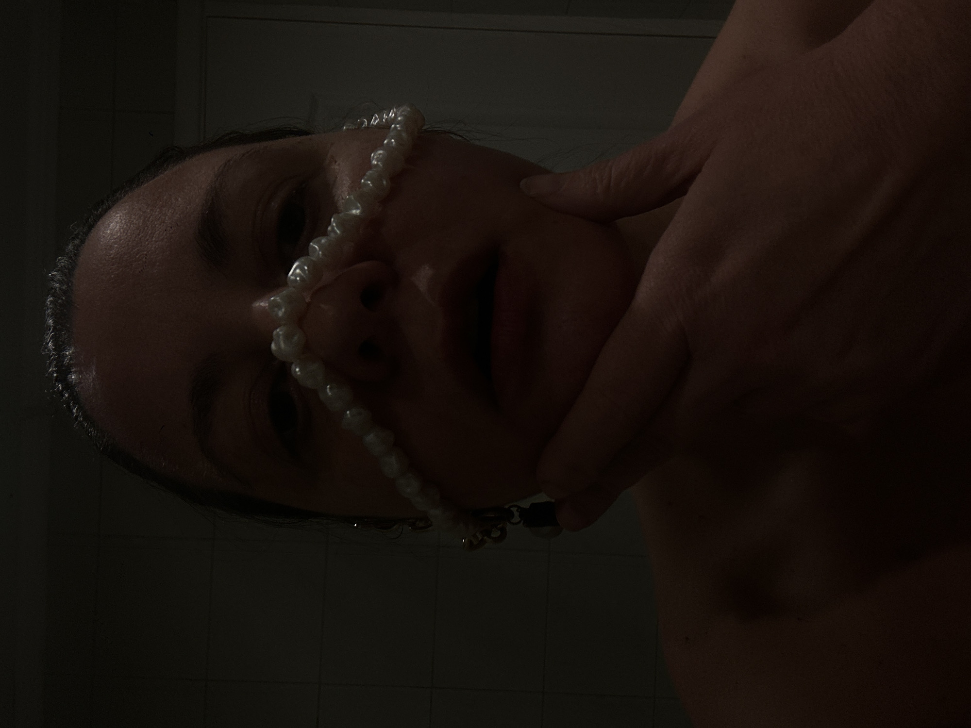 A low-angle close-up photograph depicts a person's face adorned with a string of white pearls draped across the nose. The person's dark hair is visible. A human hand touches the person's cheek. The image has a predominantly dark and grainy texture. The location is not discernible as Riga, Latvia. The environment is not identifiable.
