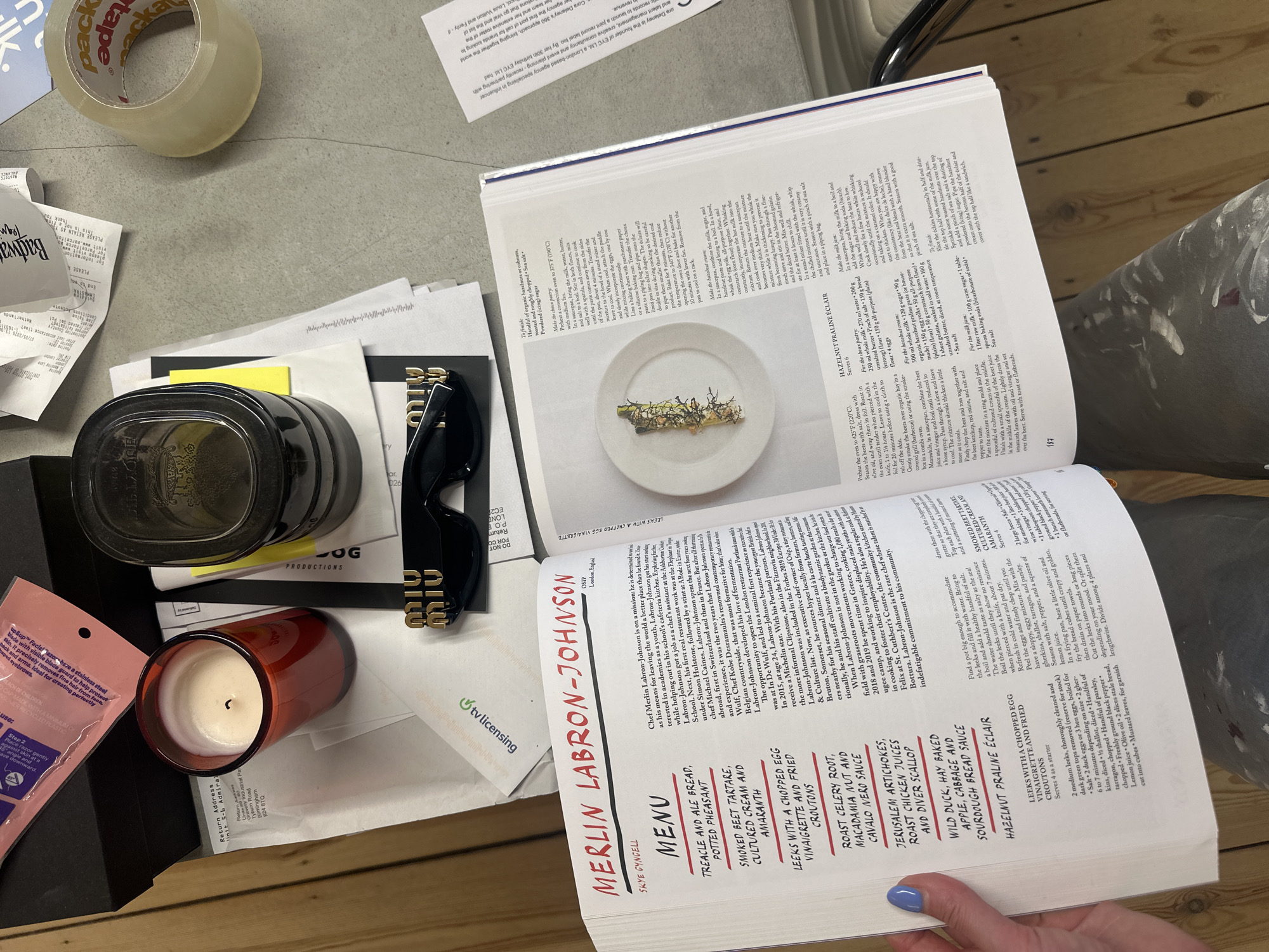 The image depicts a cluttered desk or table with various items scattered around a cookbook opened to a recipe for "Merlin Labron-Johnson". On the left side of the image, there are receipts, a black container, a red candle in a glass jar, a pink package, and black sunglasses. At the top, there's packing tape. The cookbook lies open, with a recipe for "Hazlenut Praline Eclair" visible. In the lower right, a hand with blue nail polish holds the cookbook open.